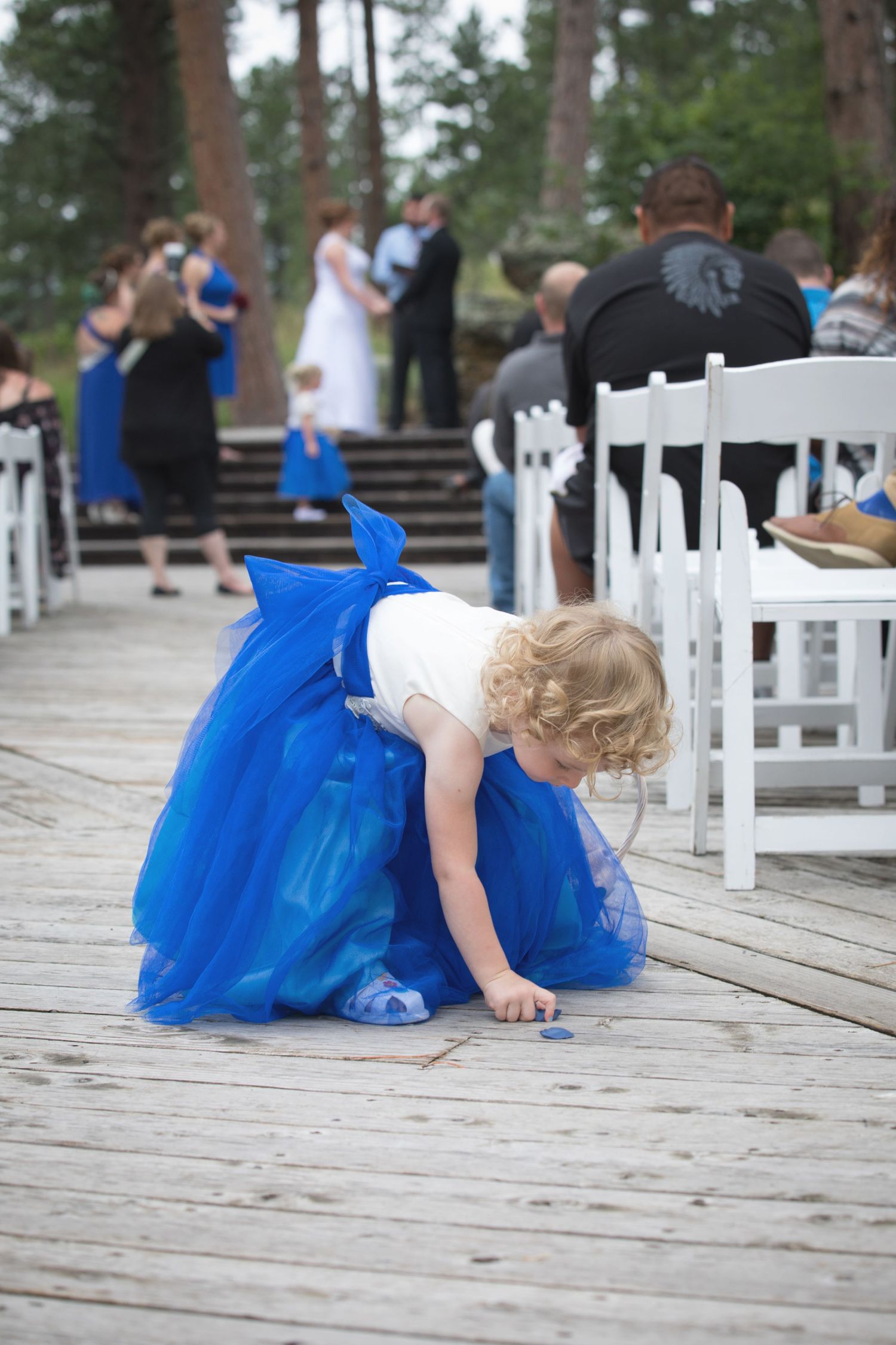 Young flower girl in blue tulle dress bending down on wooden deck at outdoor wedding ceremony.