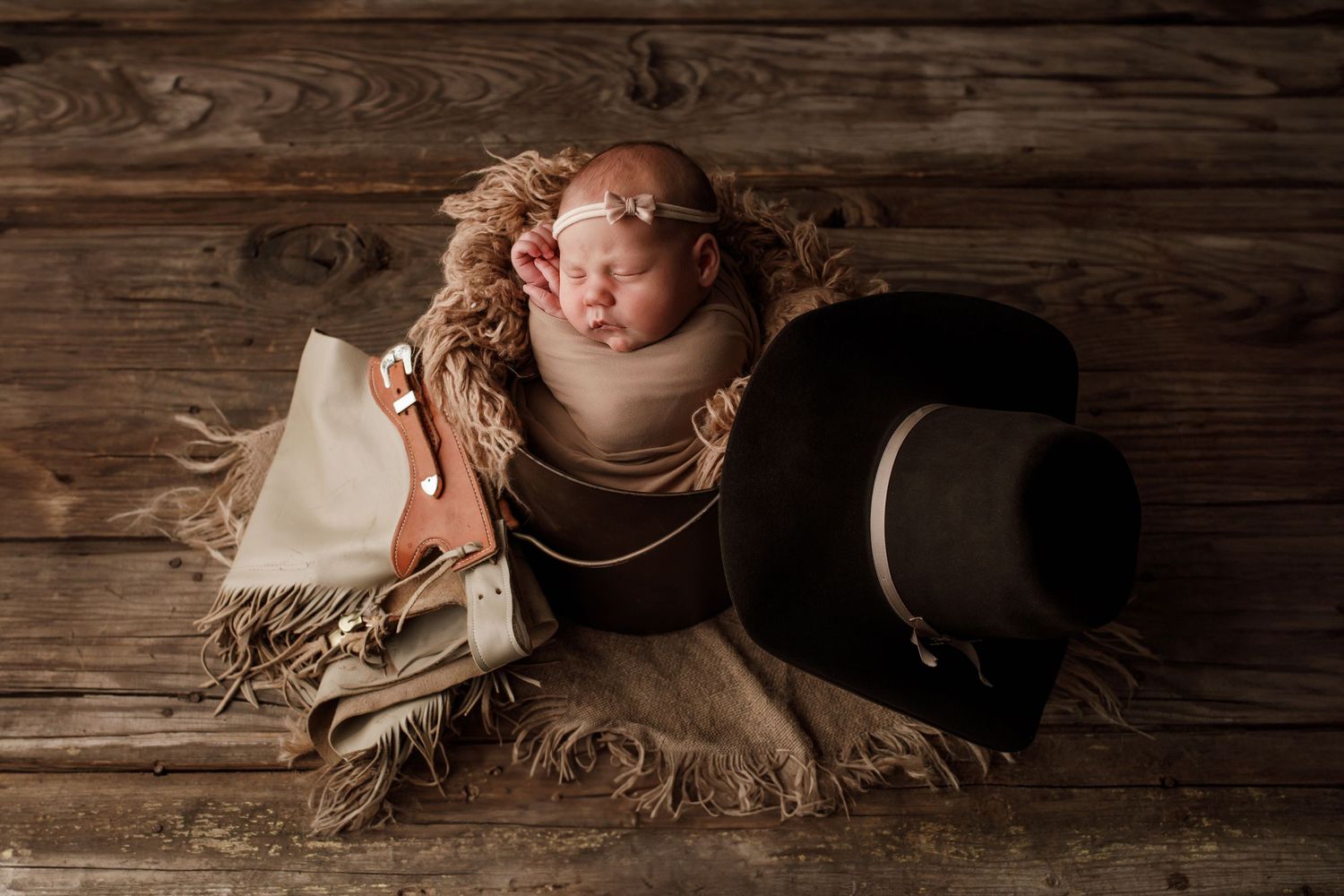 Sleeping swaddled newborn baby girl posed with cowboy hat and western props on wood.