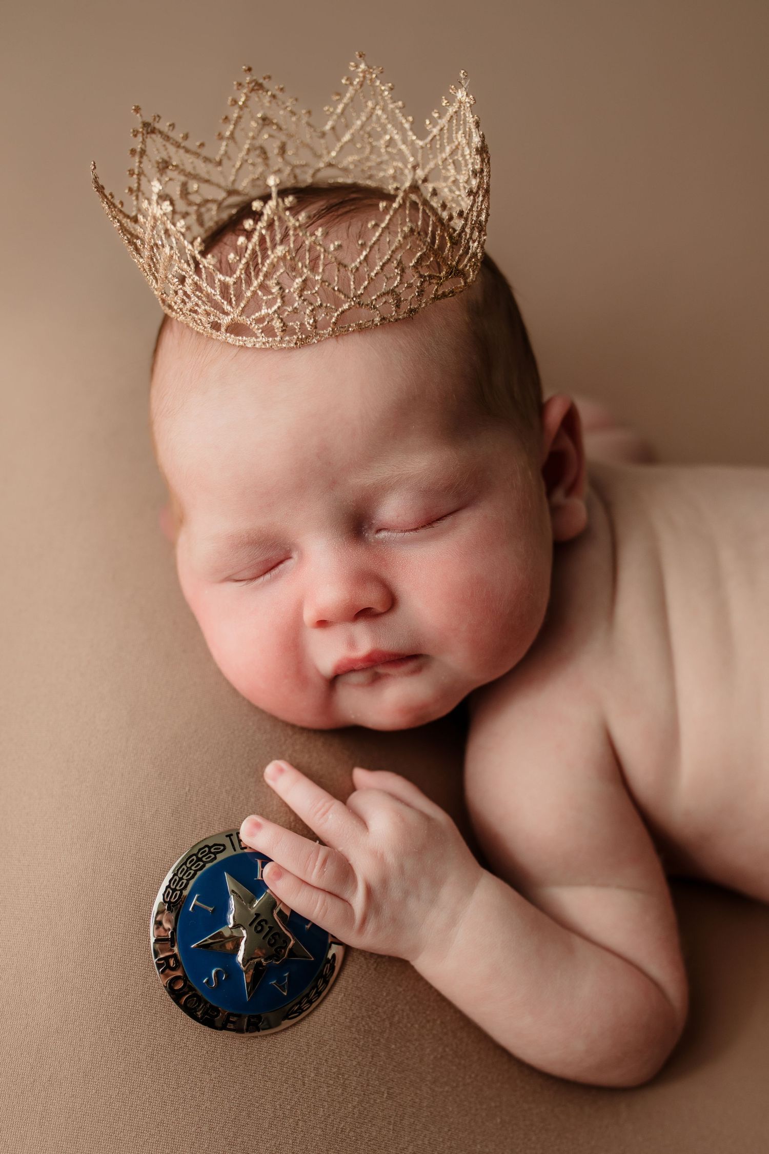 Sleeping newborn baby wearing gold lace crown holding decorative badge on beige background.