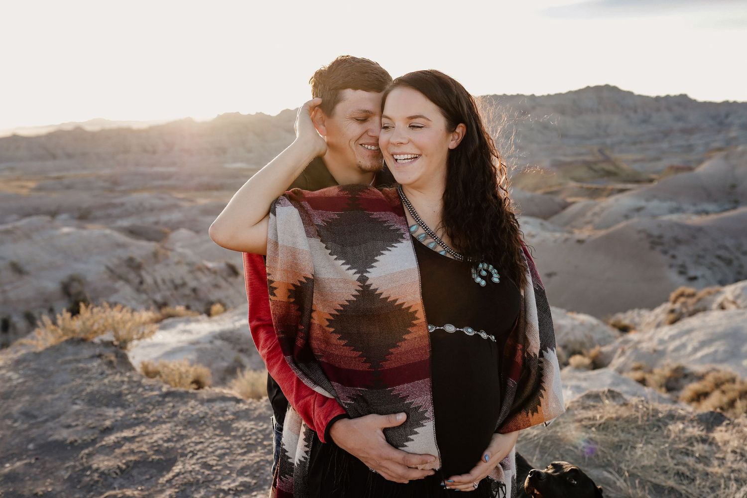 Couple embracing at sunset in badlands, man in aztec blanket wrap holding pregnant woman's belly.