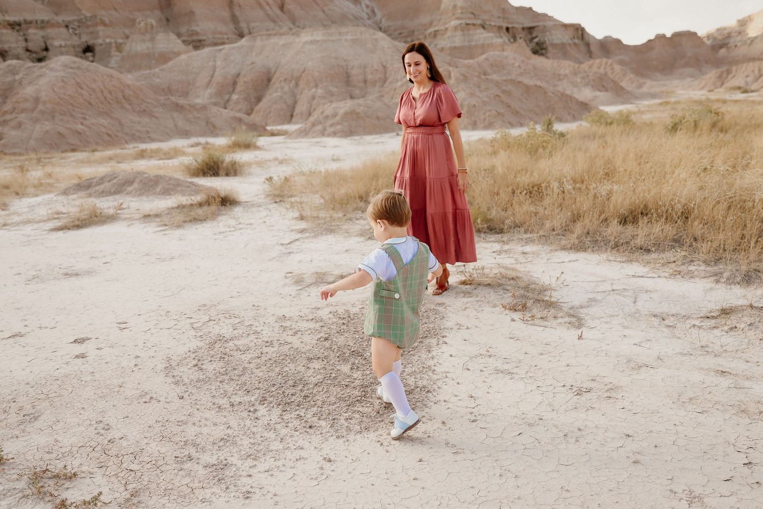 Young toddler in green romper running toward mother in pink dress in scenic desert landscape.
