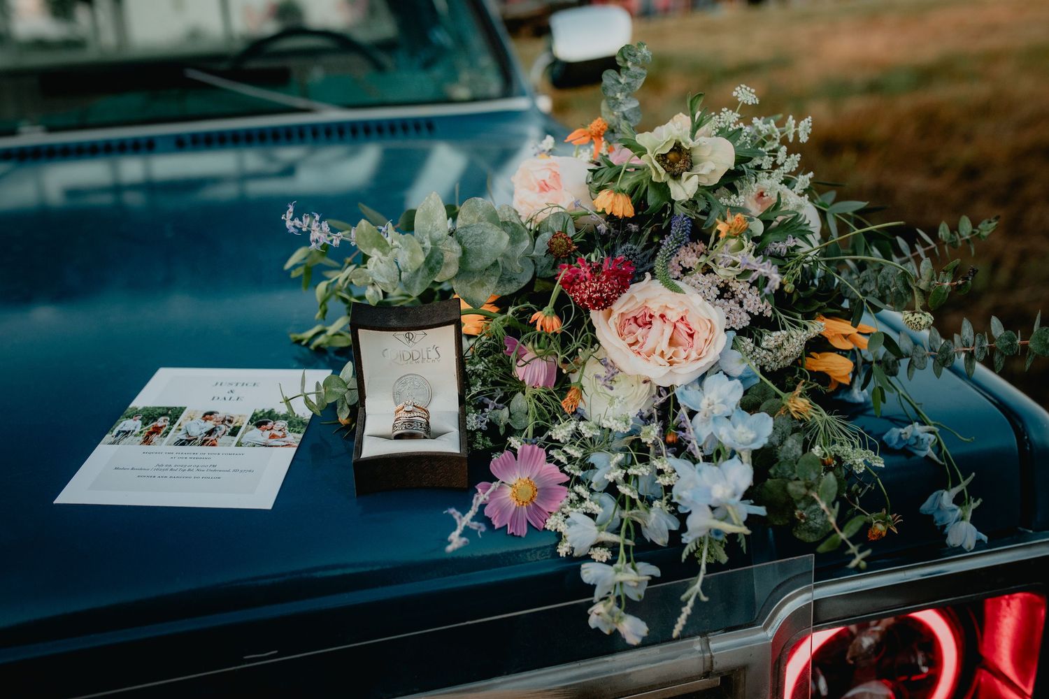 Wedding bouquet with pink roses and wildflowers resting on a vintage blue car hood with ring box and invitation.