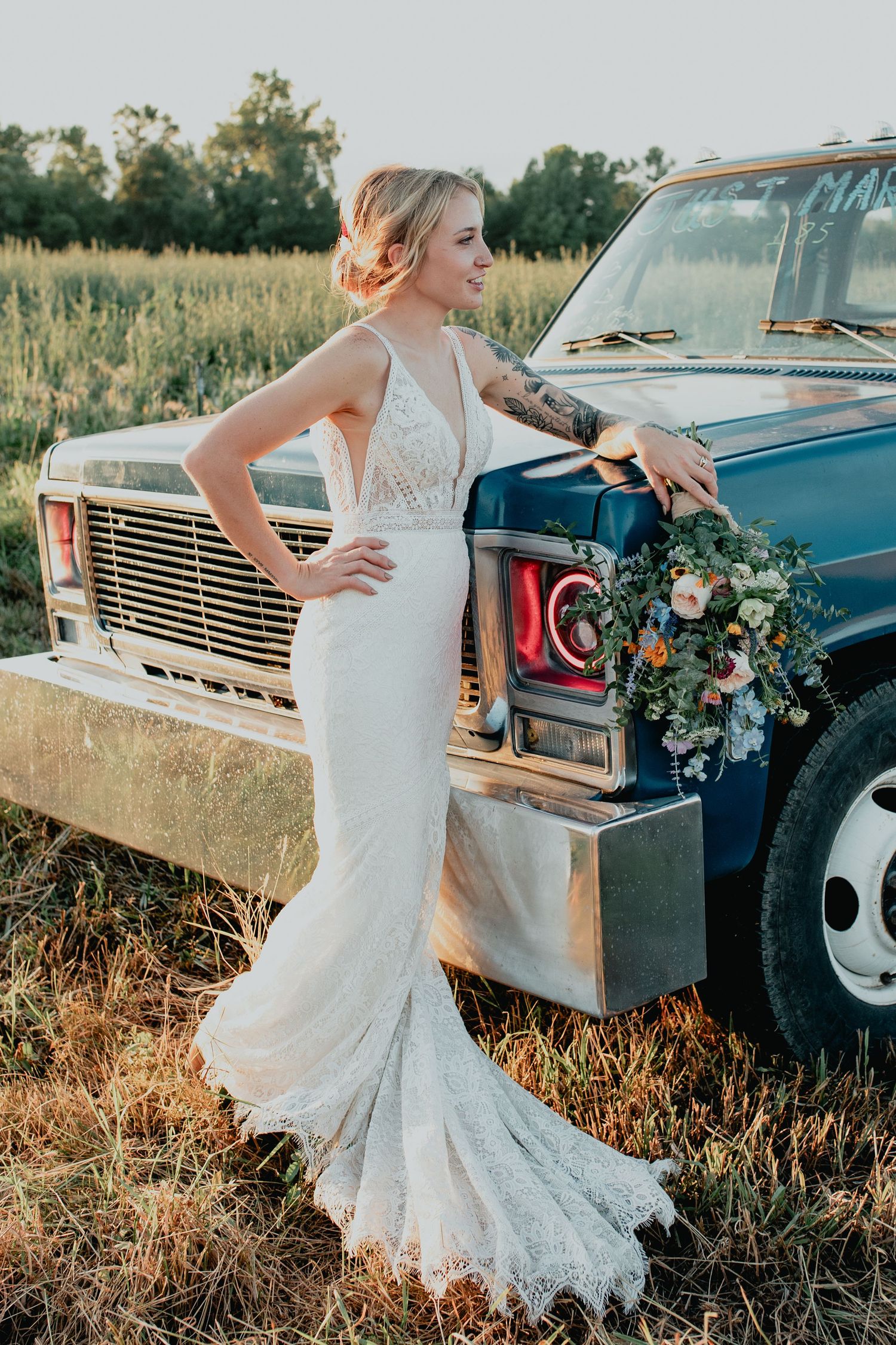 Bride in lace mermaid gown leans against vintage blue truck holding floral bouquet in golden hour field.