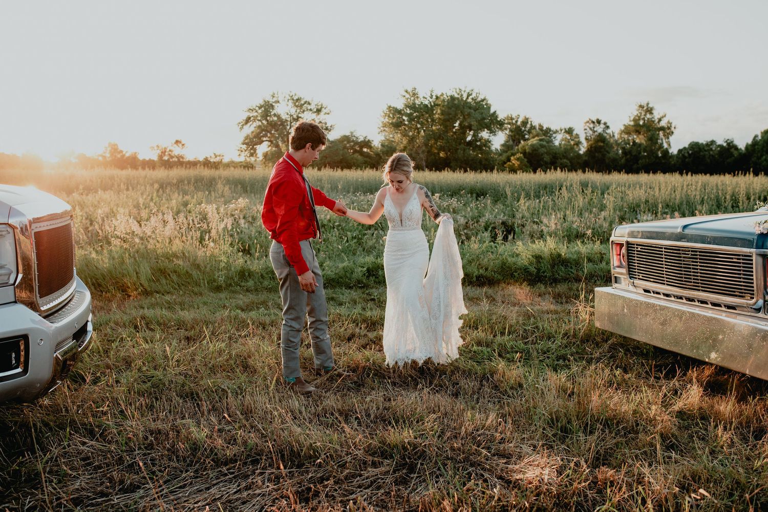 Bride and groom holding hands at golden hour between vintage trucks in a rustic field setting.
