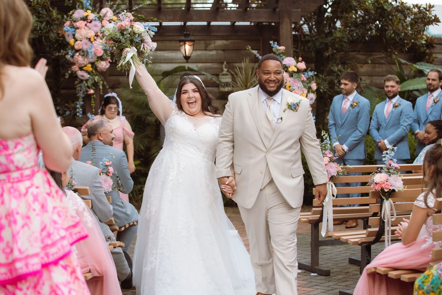Joyful newlyweds walk down the aisle, bride raising bouquet, guests in pink and blue attire at outdoor wedding.
