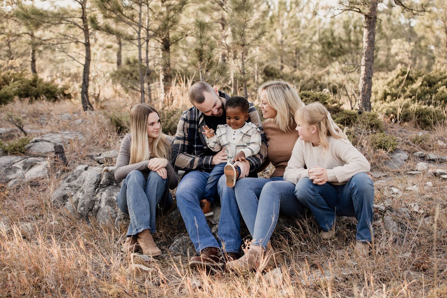 Family of five sitting on rocks in a forest, sharing a joyful moment with a laughing toddler outdoors.