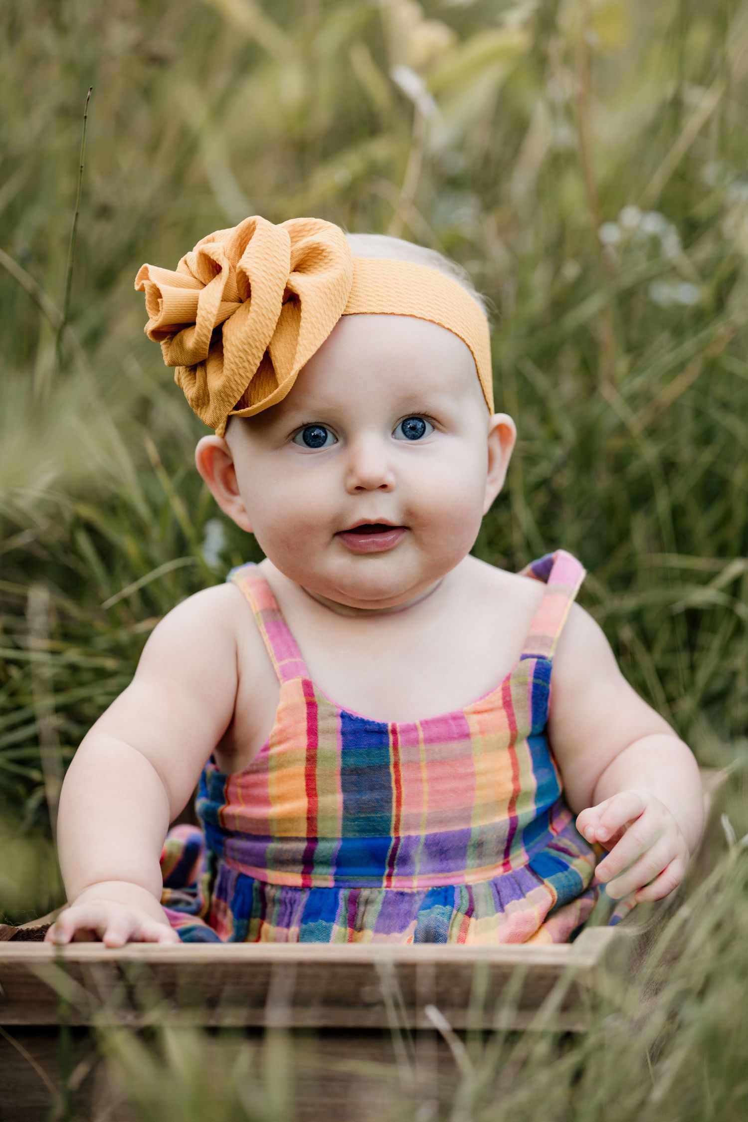 Cute baby girl with blue eyes wearing a colorful plaid dress and gold bow headband outdoors.