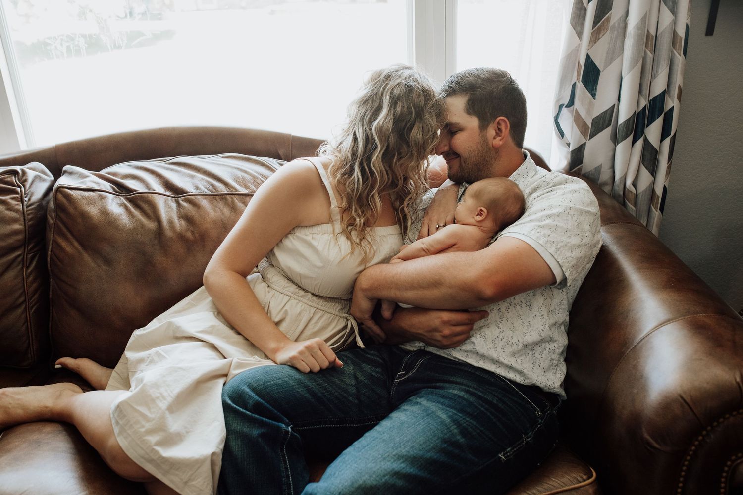 New parents cuddling together on leather couch holding their newborn baby at home.
