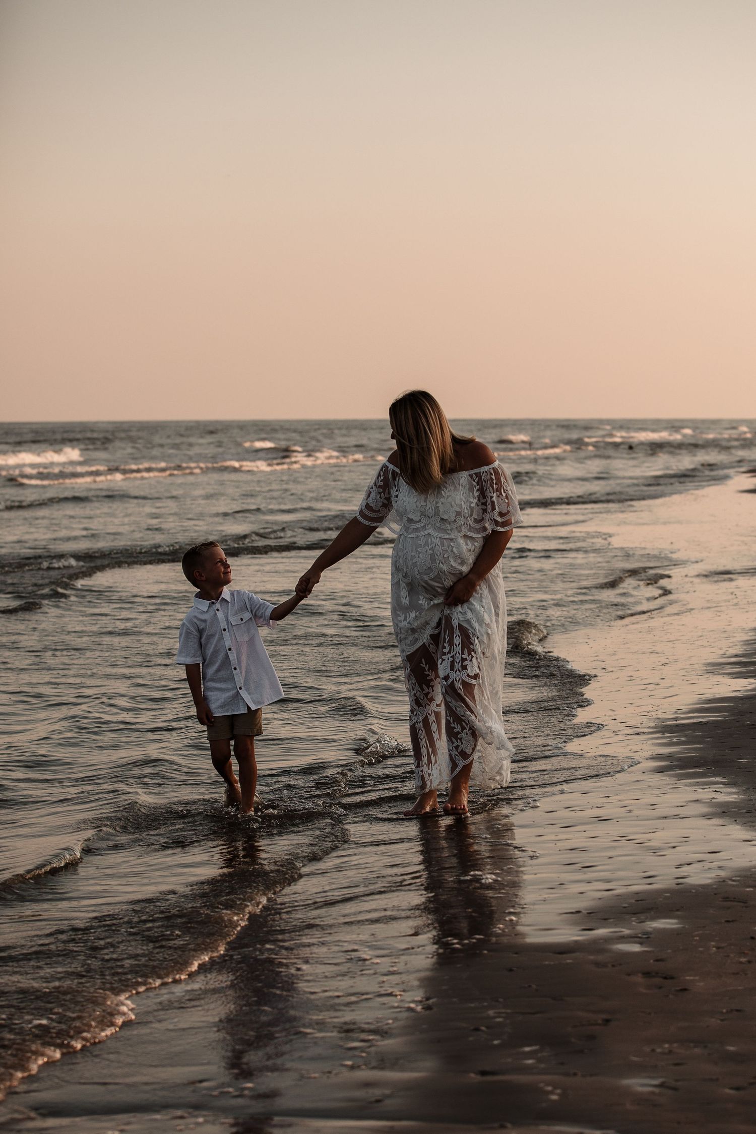 Pregnant woman in white lace dress holding hands with young boy walking along ocean shoreline at dusk.