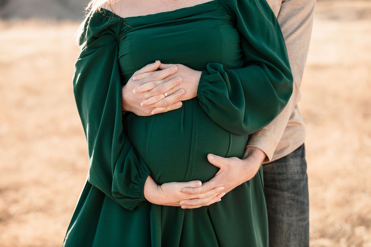 Someone wearing a green dress holds their pregnant belly while being embraced from behind in a warm outdoor setting.
