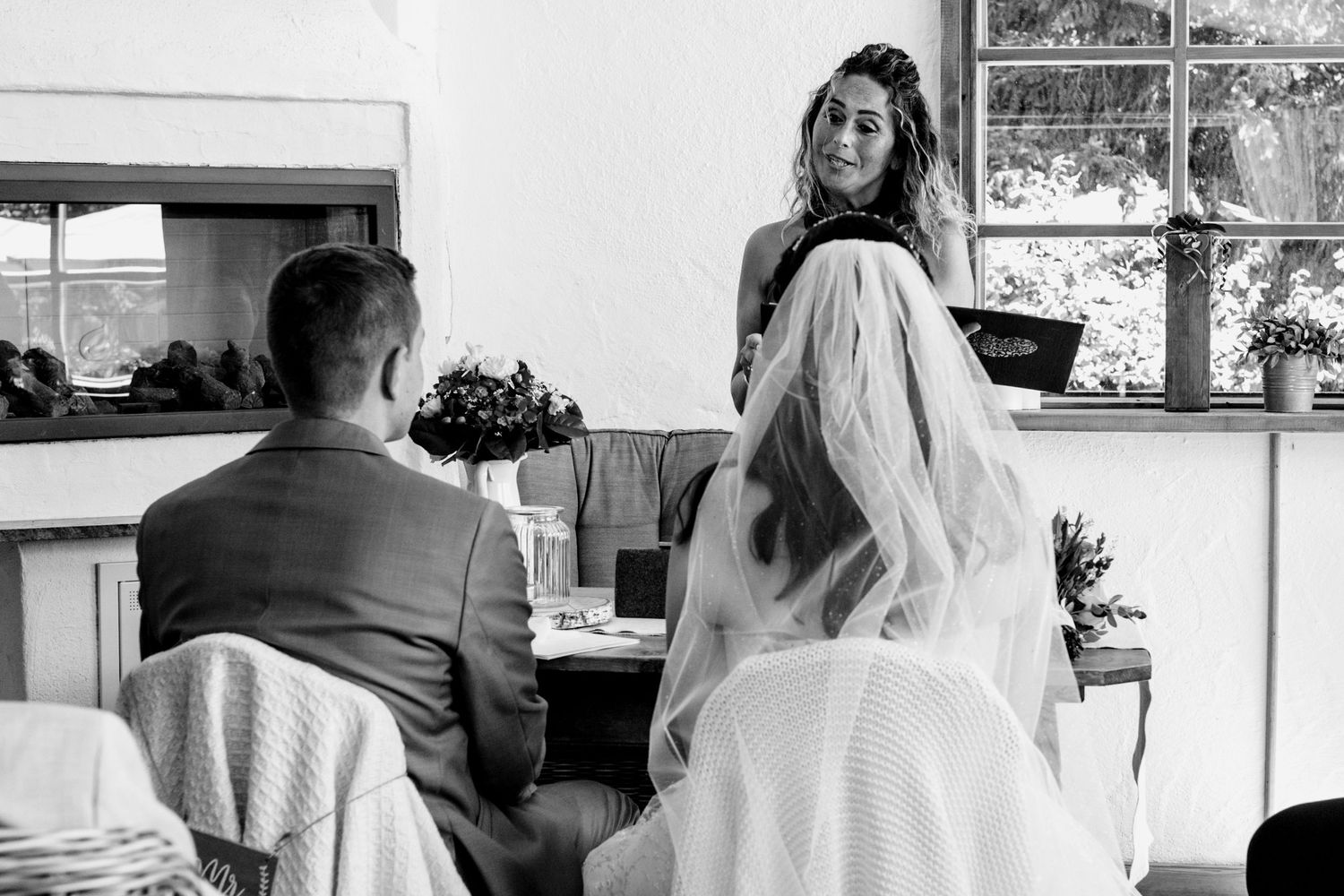 Black and white wedding ceremony photo showing bride and groom seated before officiant holding book.