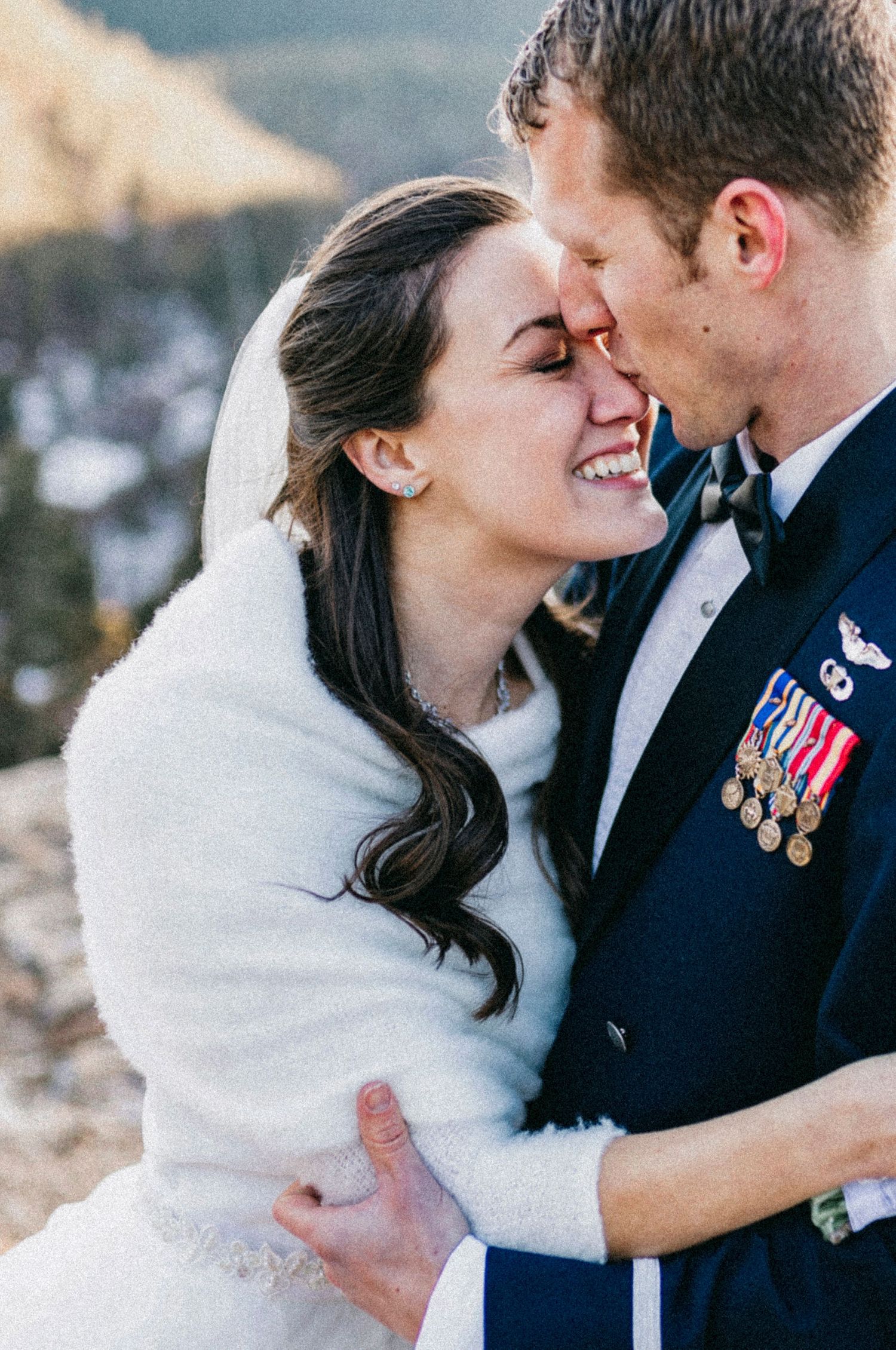 Military groom in uniform with medals embraces smiling bride in white fur wrap outdoors at winter mountain wedding.