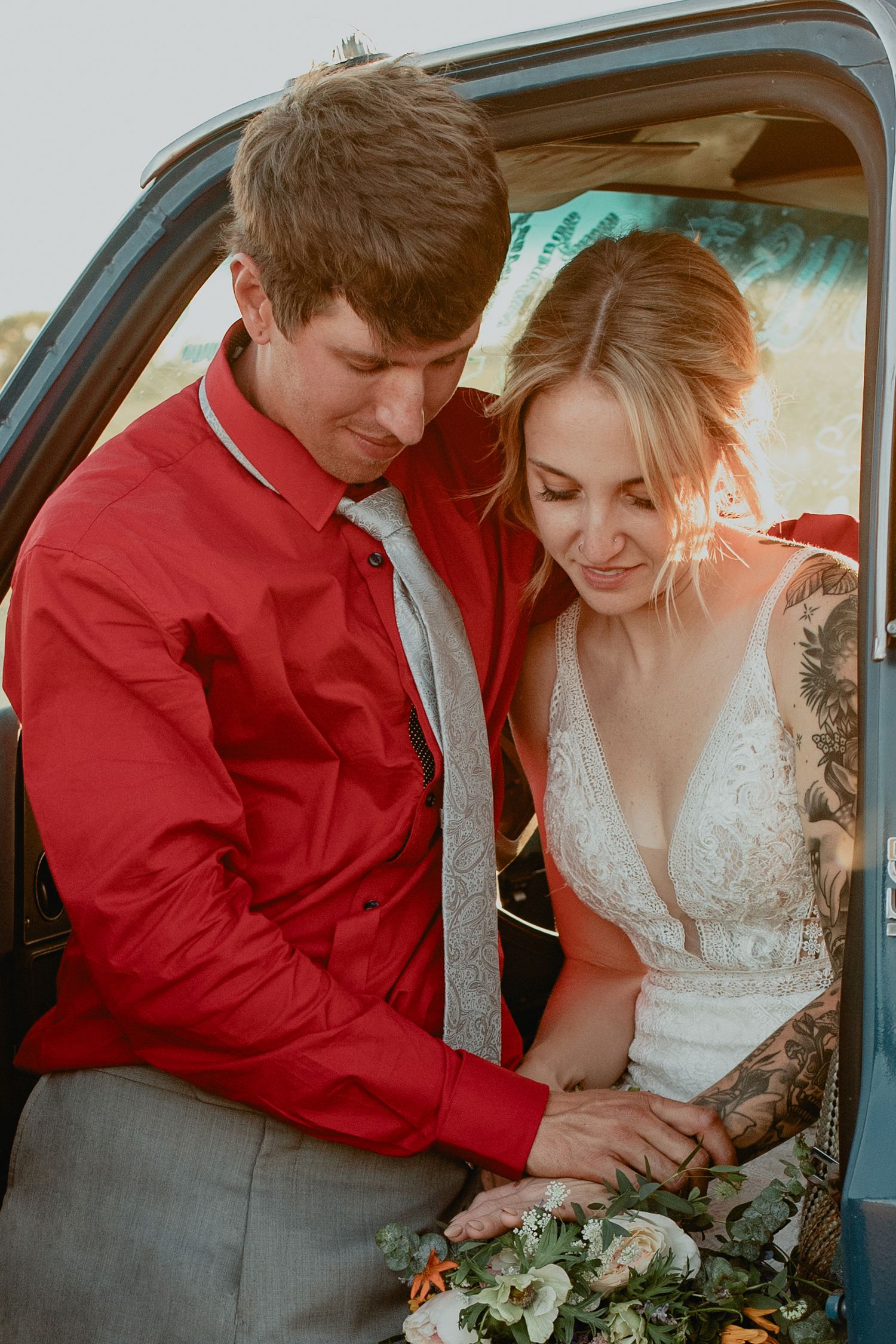 Bride in lace gown and groom in red shirt share a tender moment with a bouquet beside a vintage car.