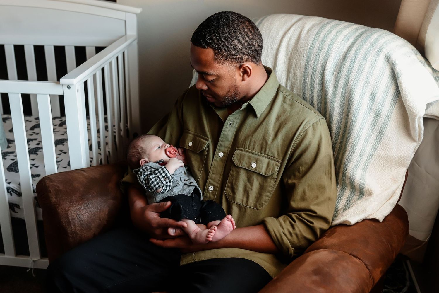 Father in olive green shirt sits in leather nursery chair lovingly gazing at newborn baby in his arms.