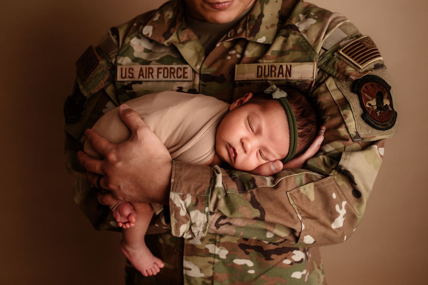 US Air Force soldier in camouflage uniform gently holding sleeping swaddled newborn baby.