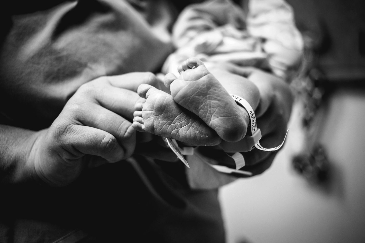 Close-up black and white photo of a parent holding a newborn baby's tiny feet with ID bracelet.