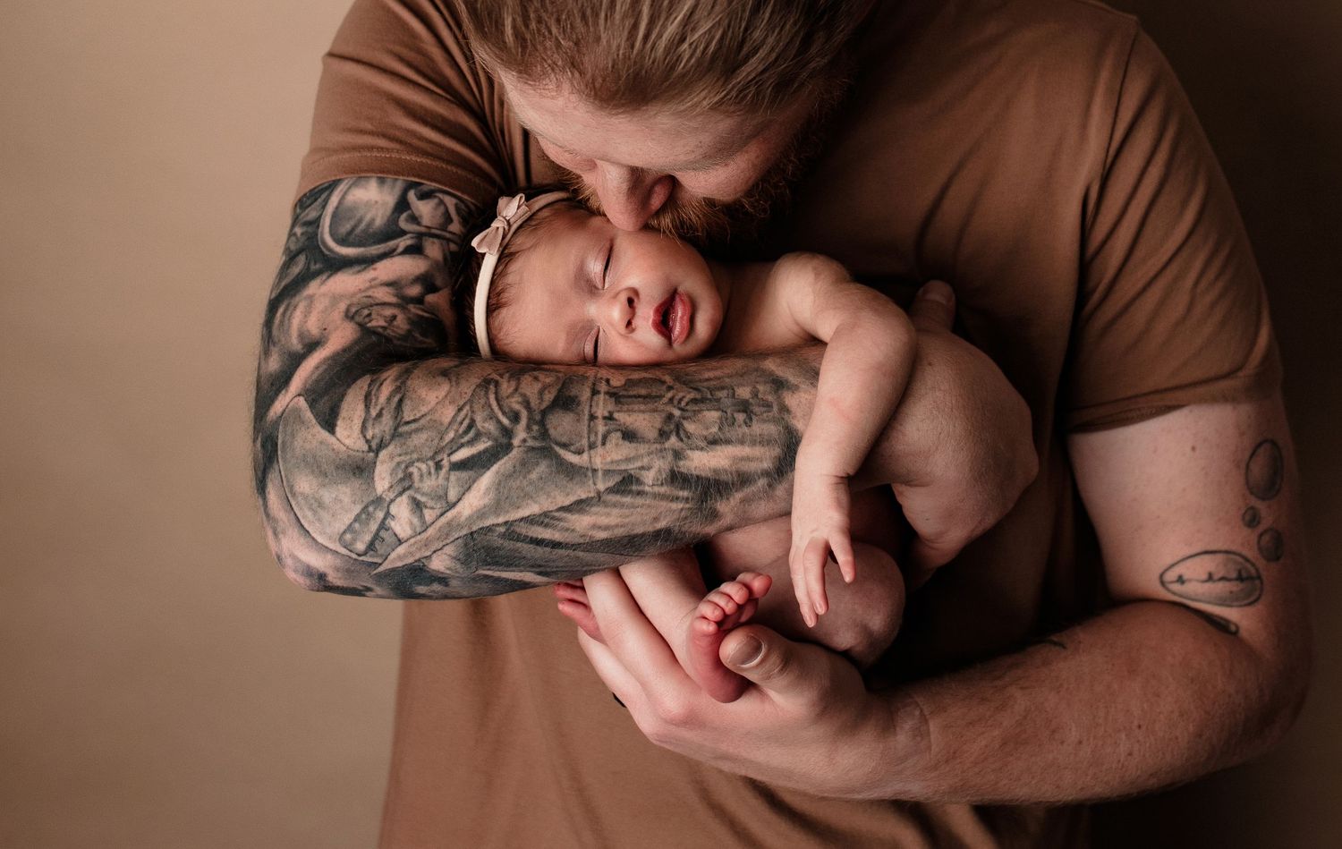 Tattooed father kissing sleeping newborn baby held gently in his arms.