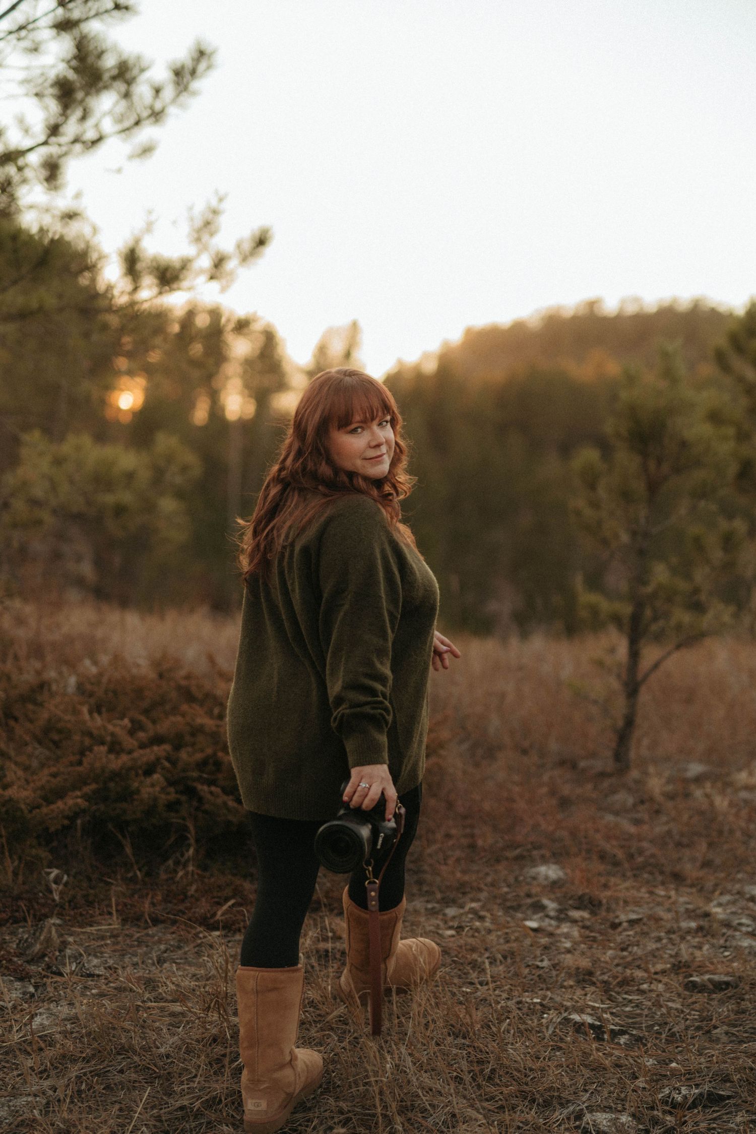 Redhead female photographer holding a camera smiling at sunset in a golden autumn field.