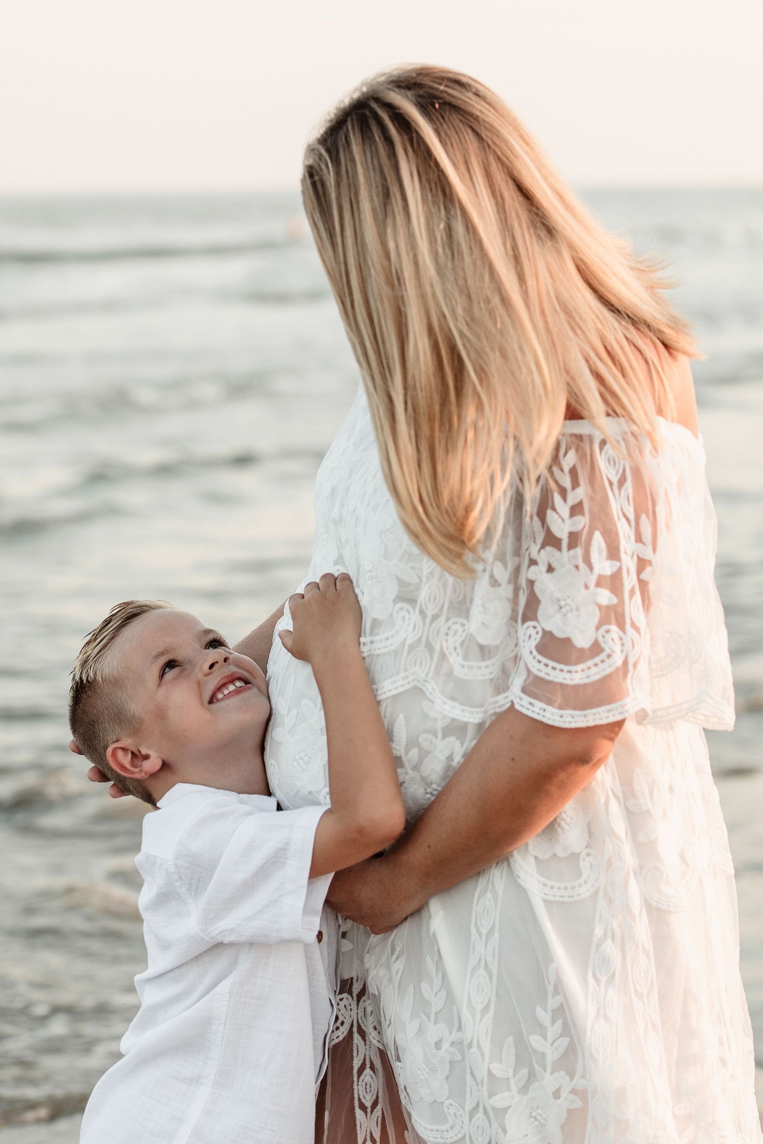 Young smiling boy hugging pregnant mother in white lace dress near ocean at golden hour.