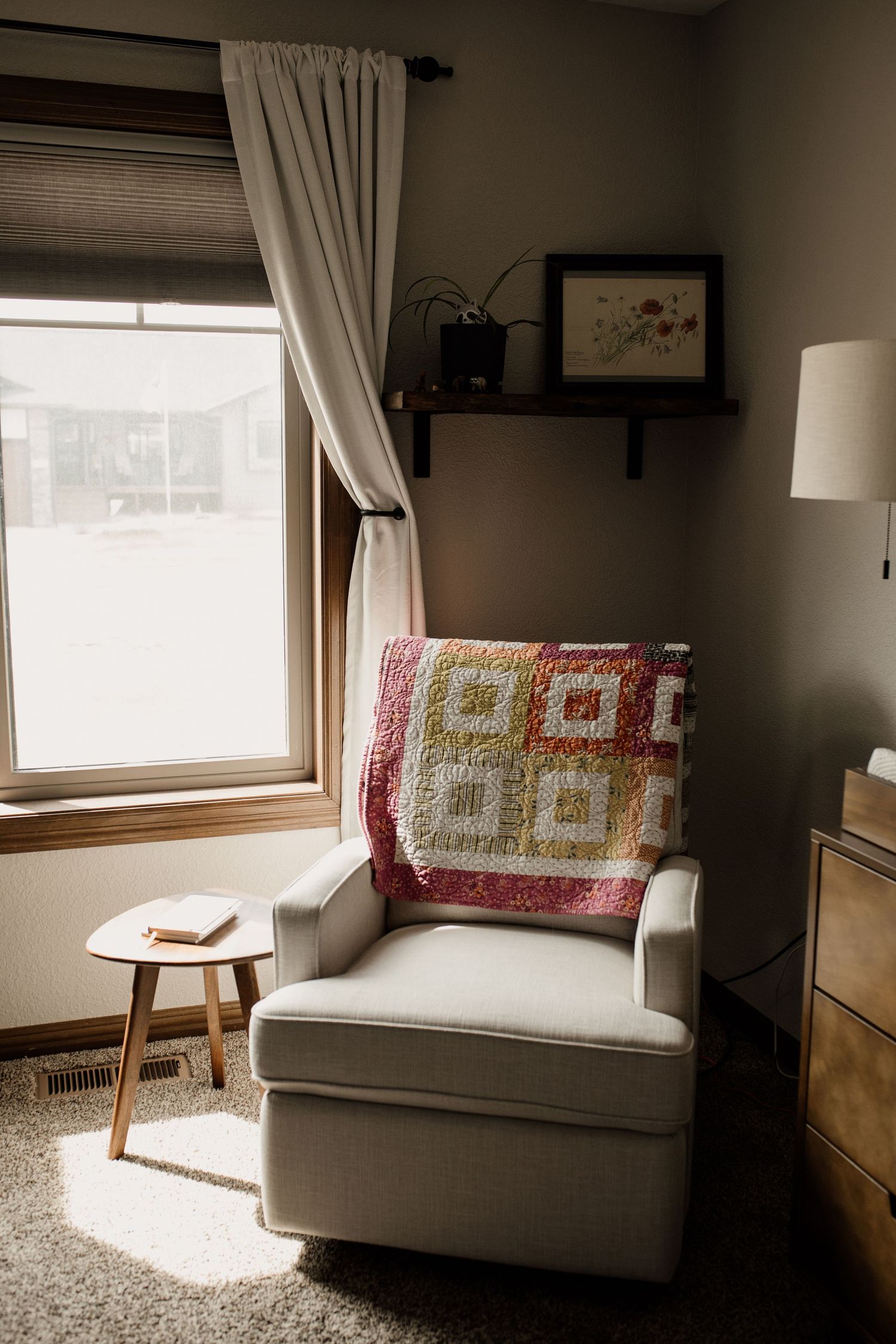 Cozy nursery corner with gray armchair, colorful quilt, and natural light from a curtained window.