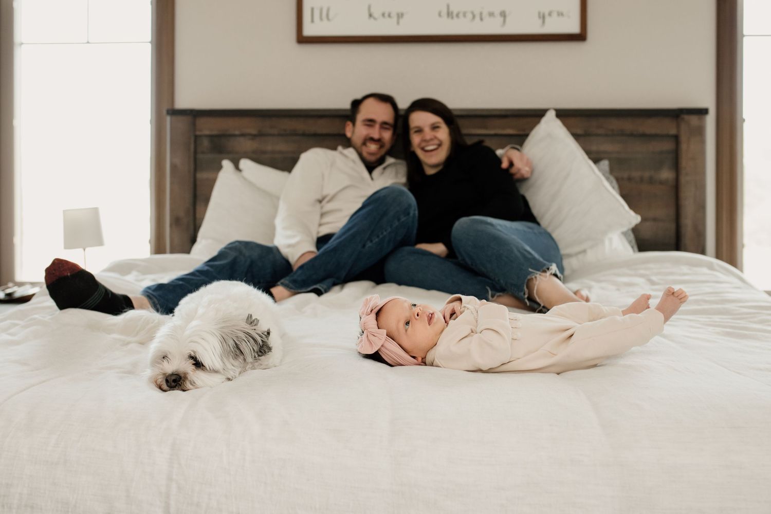 Happy couple relaxing on a white bed with their fluffy dog and newborn baby in a pink headband.