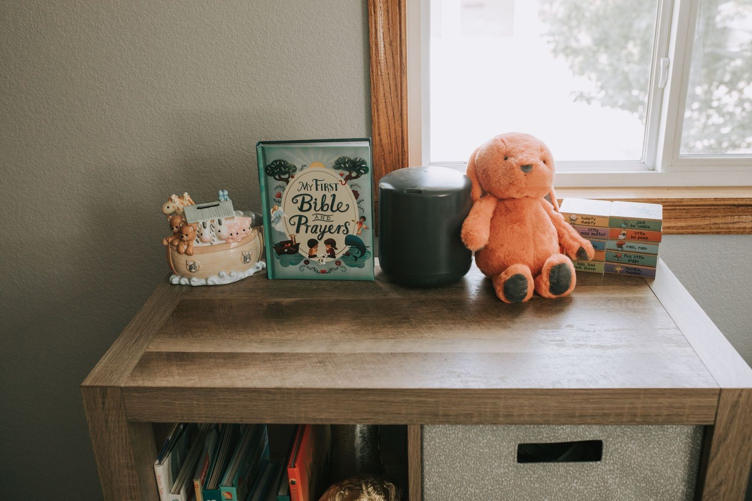 Nursery shelf decorated with a Bible prayers book, stuffed bunny, and candle by window.