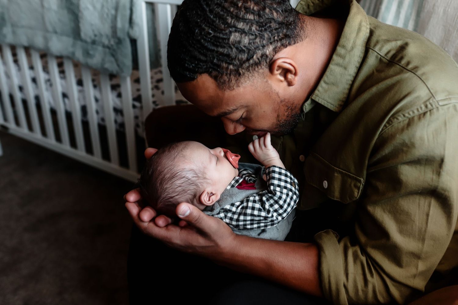 Father tenderly kisses his newborn baby while cradling the infant in his hands near a white crib.