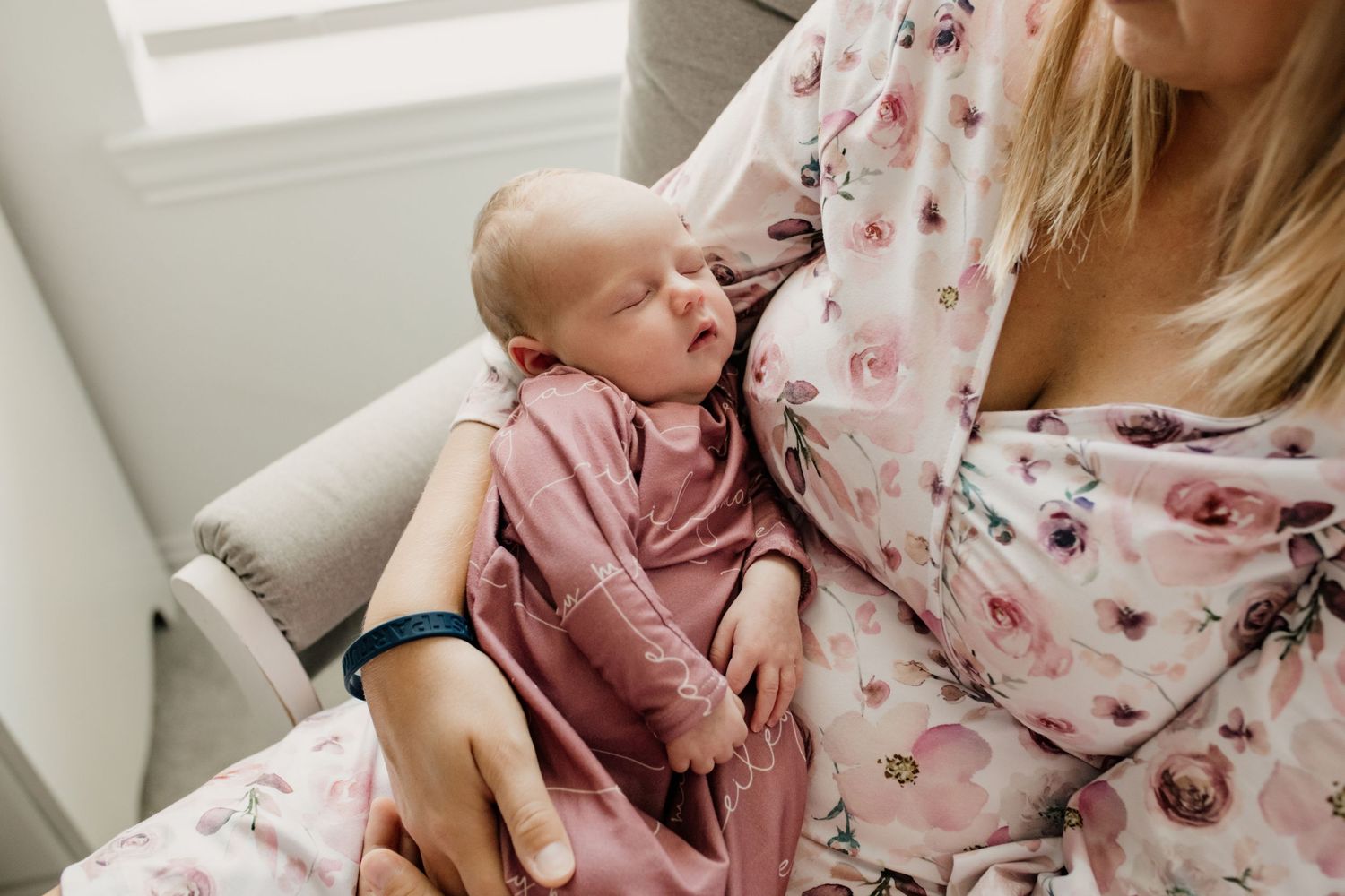 Blonde mother in floral robe cradling sleeping newborn baby dressed in dusty rose swaddle.