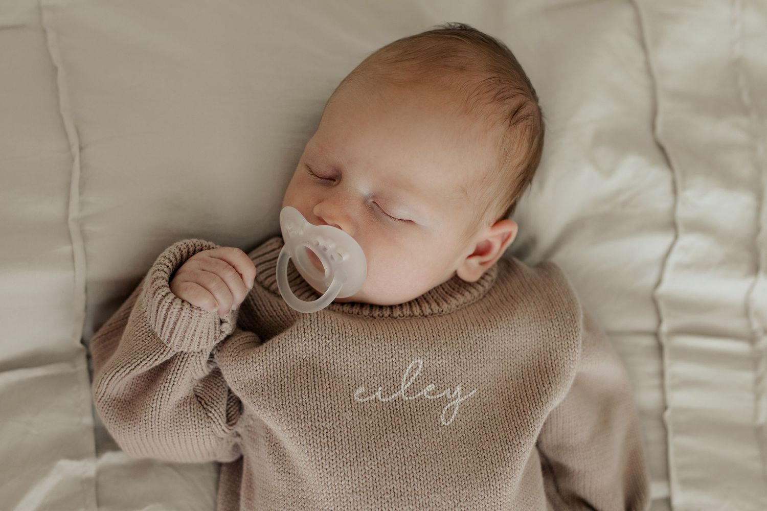 Sleeping newborn baby in a beige knit sweater with name Ciley embroidered, holding a pacifier.