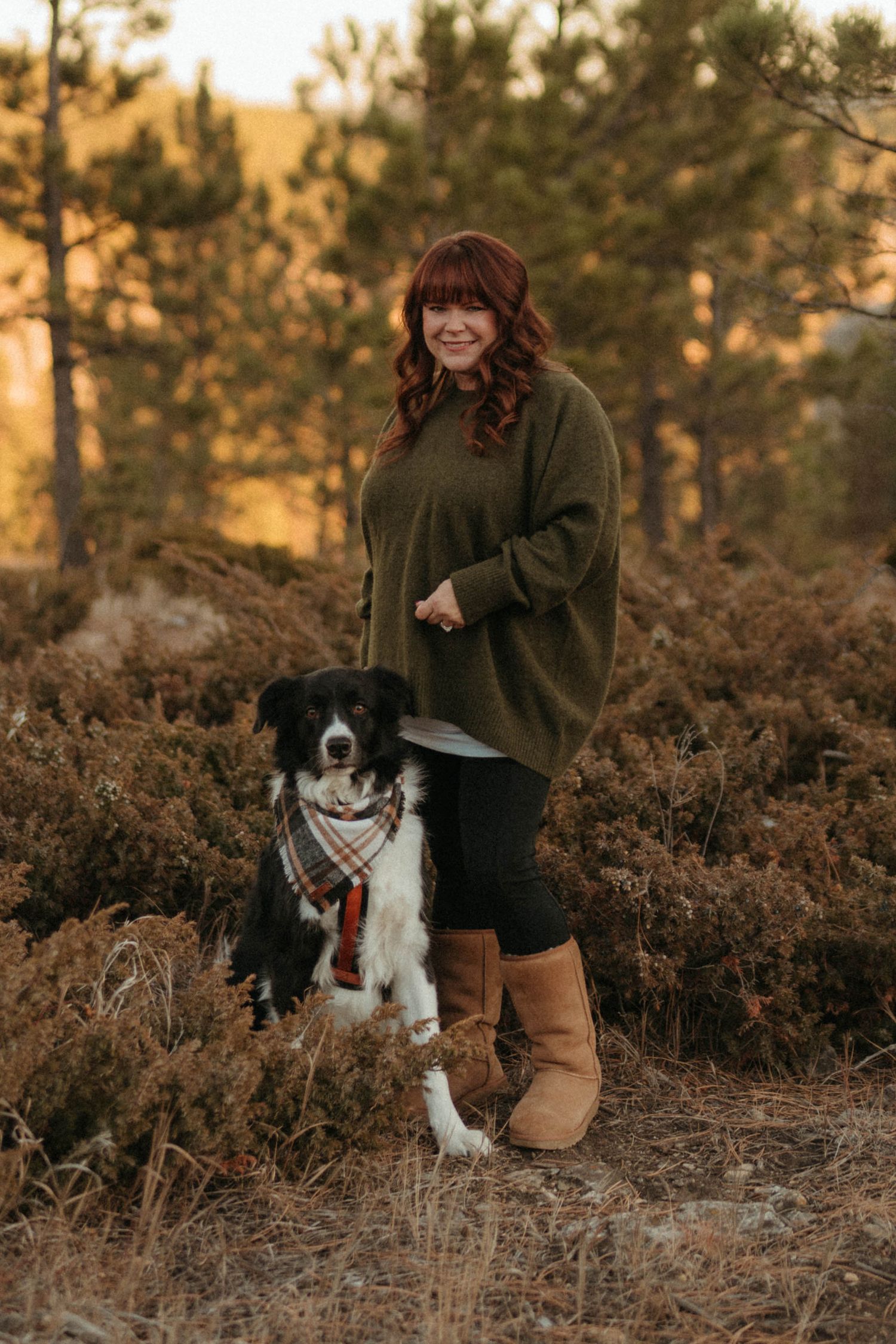 Smiling woman in green oversized sweater and UGG boots posing with her Border Collie in autumn woodland.