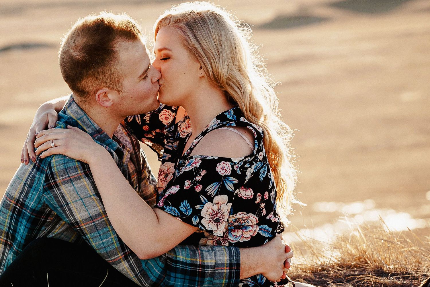 A couple shares a romantic kiss while embracing outdoors in a sandy landscape, woman in floral dress and man in plaid shirt.