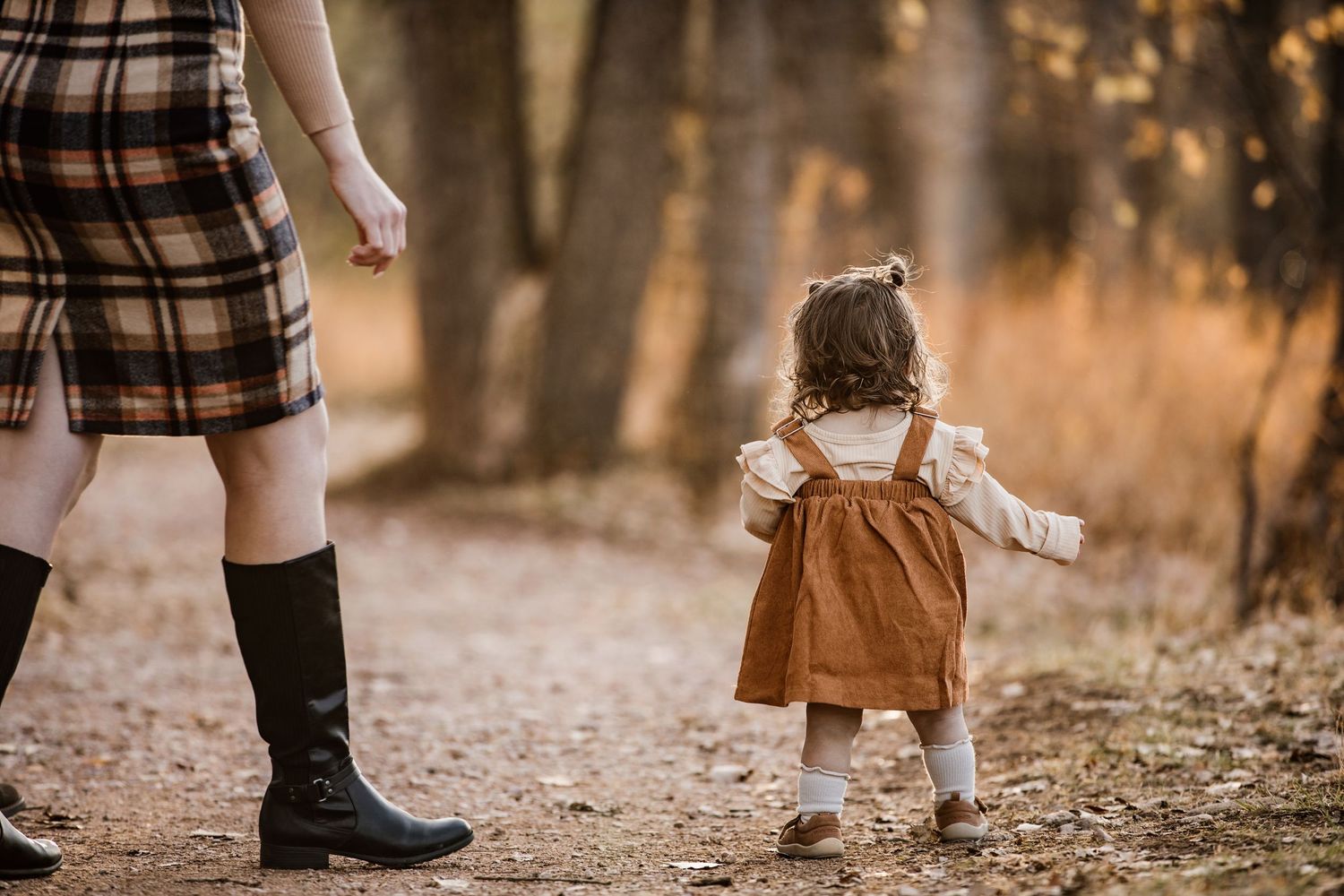 Toddler girl in brown dress walks with mother on autumn forest path.