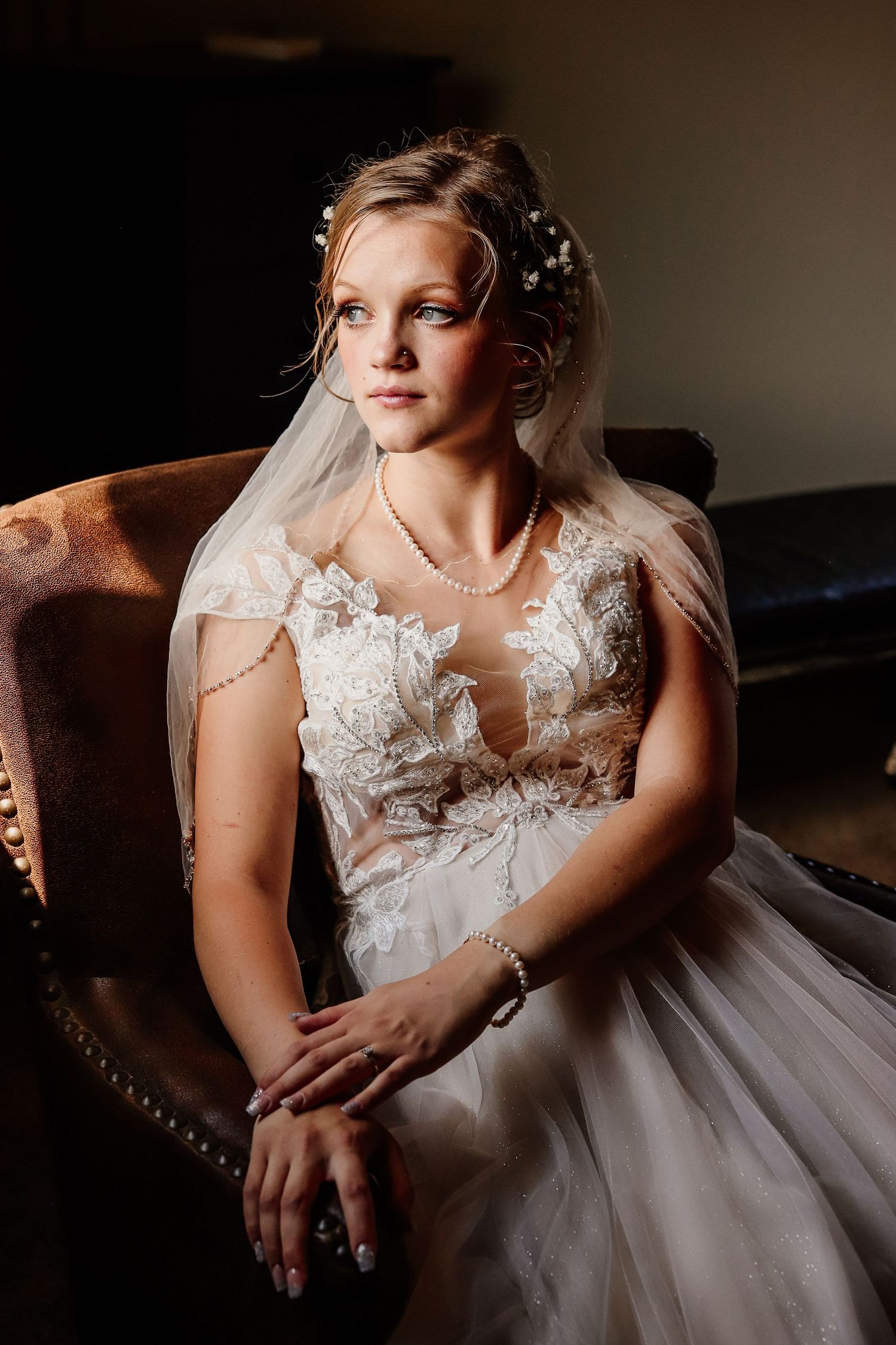 Bride in lace wedding gown and veil sits in vintage chair, wearing pearl jewelry, with soft dramatic lighting.