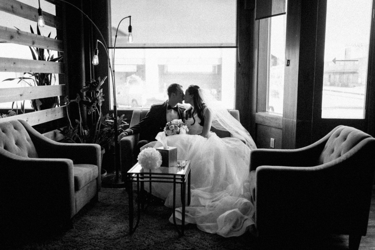 Black and white photo of a bride and groom sharing a kiss while seated on elegant chairs in a modern venue.