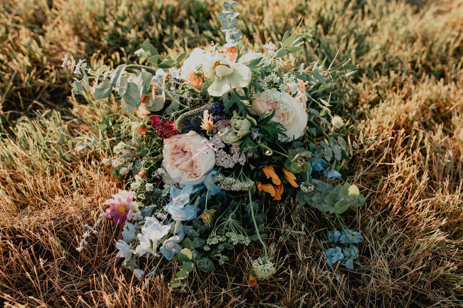 A lush bridal bouquet with pink roses, wildflowers, and greenery resting on golden grass outdoors.