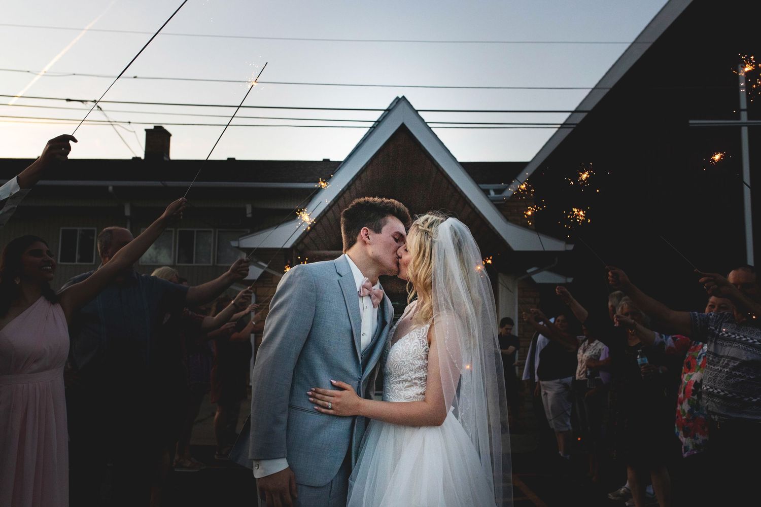Bride and groom share a romantic kiss surrounded by guests holding sparklers at twilight outdoor wedding celebration.