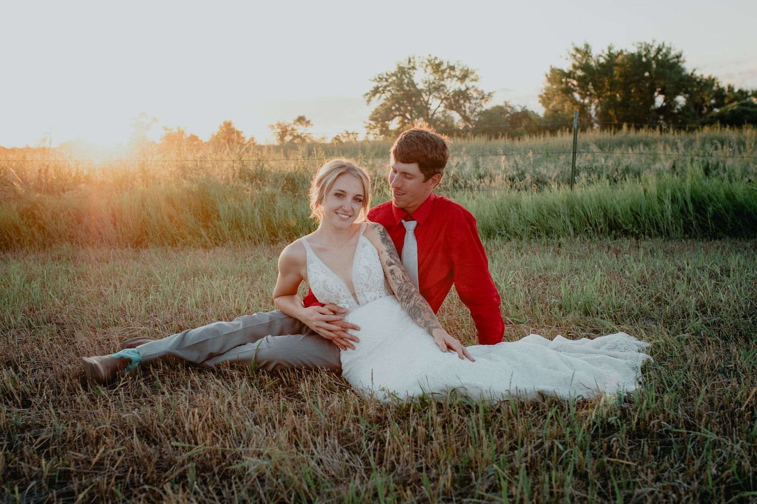 Couple sitting in golden field at sunset, bride in white gown, groom in red jacket, romantic outdoor wedding photo.