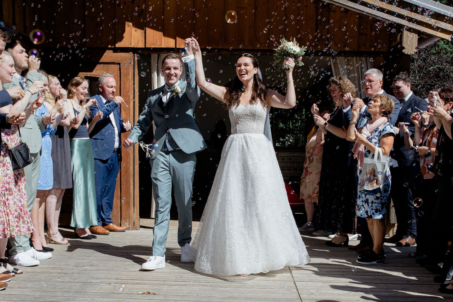 Bride and groom celebrate wedding exit with confetti shower, guests applauding outdoors at rustic venue.