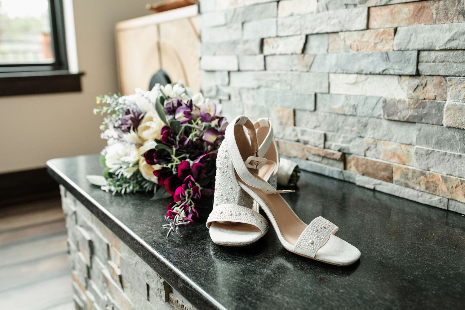 White bridal heels with crystal embellishments beside a purple and white wedding bouquet on a dark granite surface.