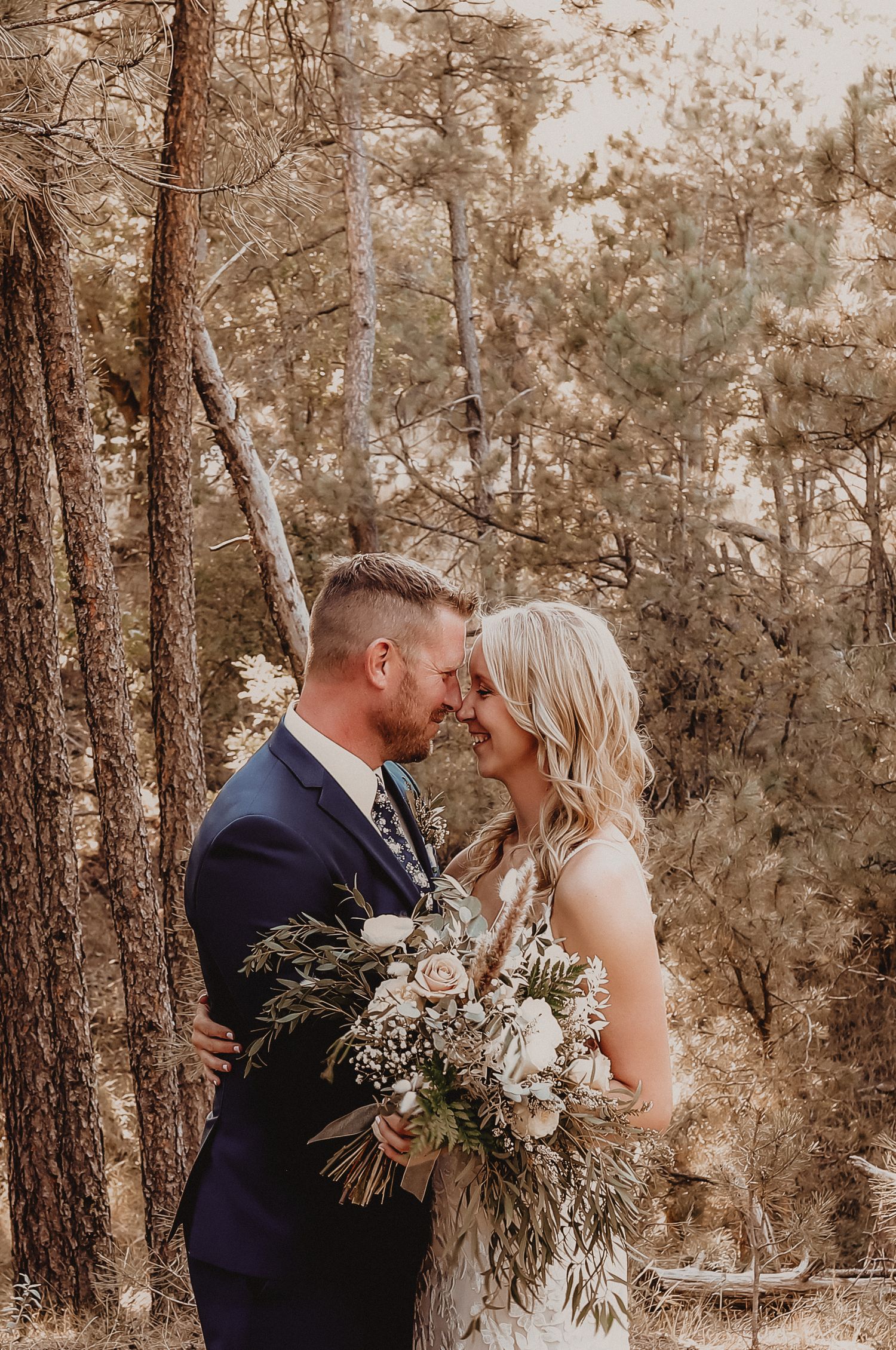 Bride and groom touching foreheads, smiling in a forest setting, bride holding a lush white and green bouquet.