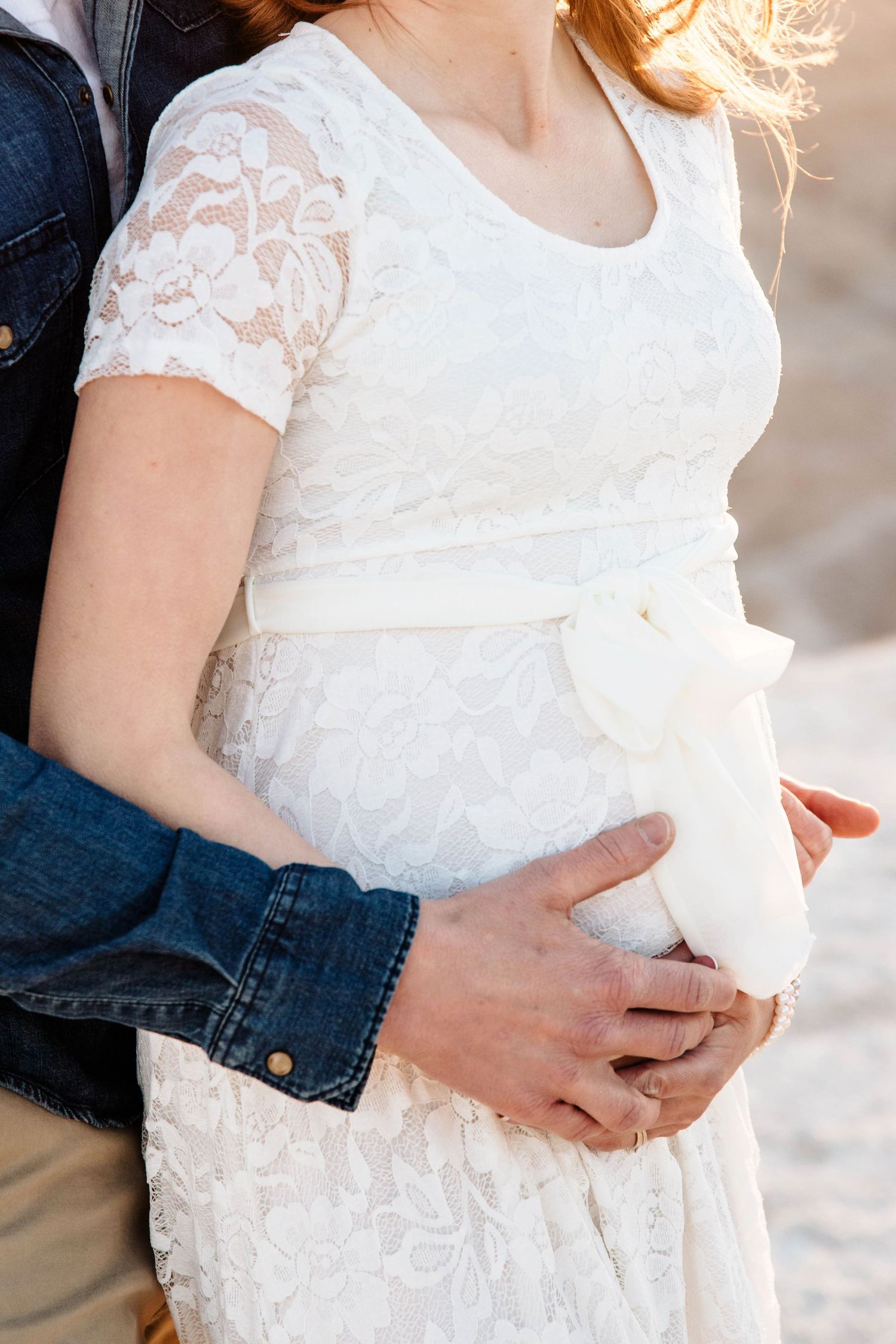 Close-up of pregnant belly in white lace dress with bow, partner embracing from behind.