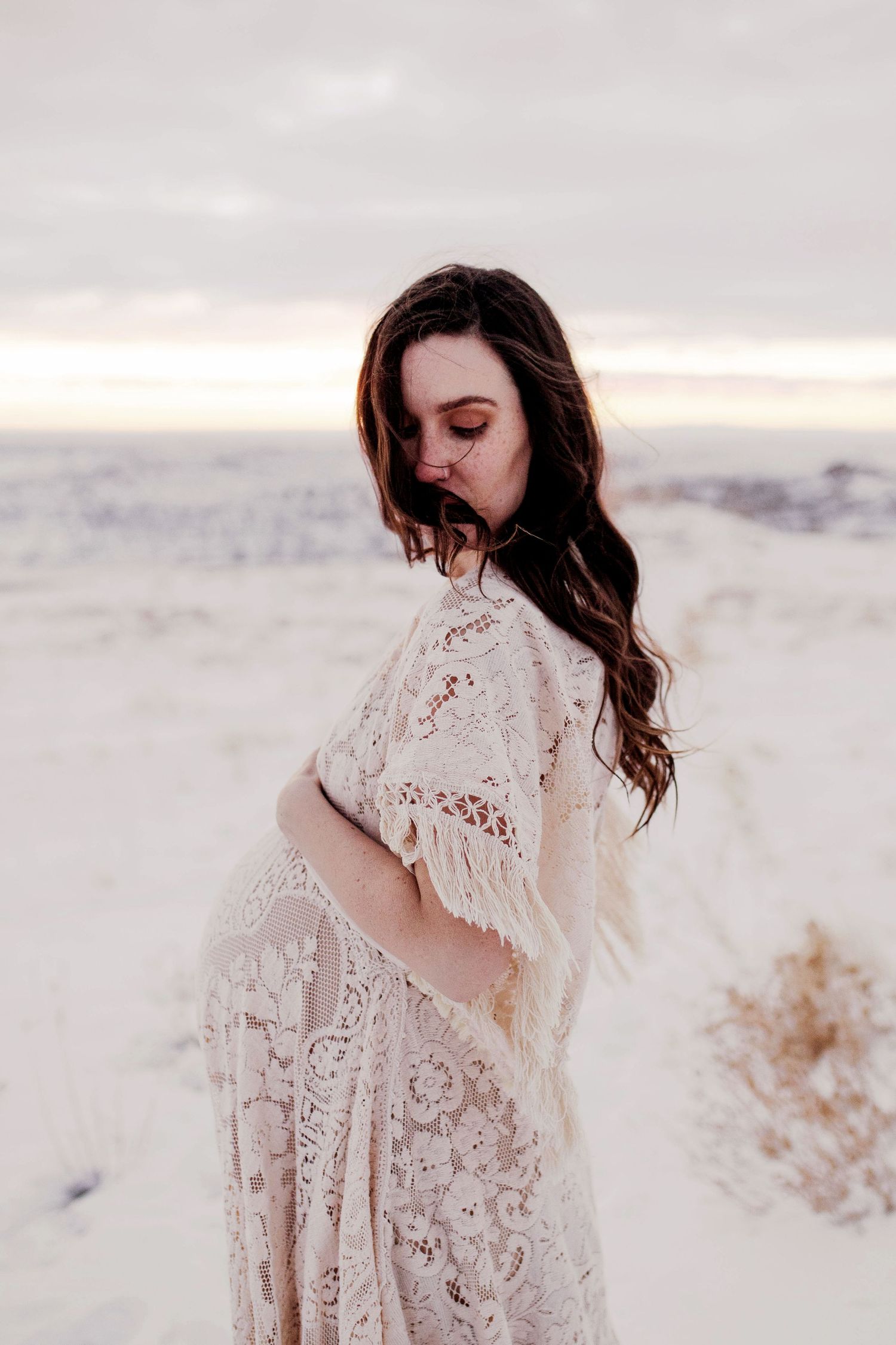 Pregnant woman in boho lace fringe wrap gazes down at bump on snowy beach at dusk.