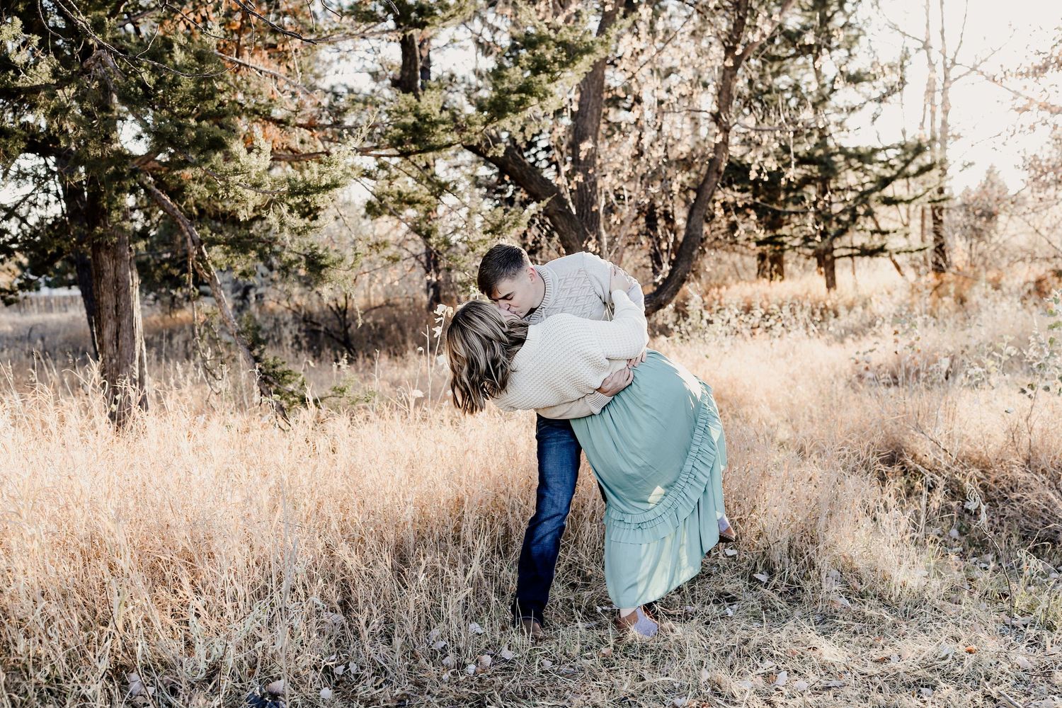 Couple sharing a romantic embrace in a golden field surrounded by pine trees during autumn.