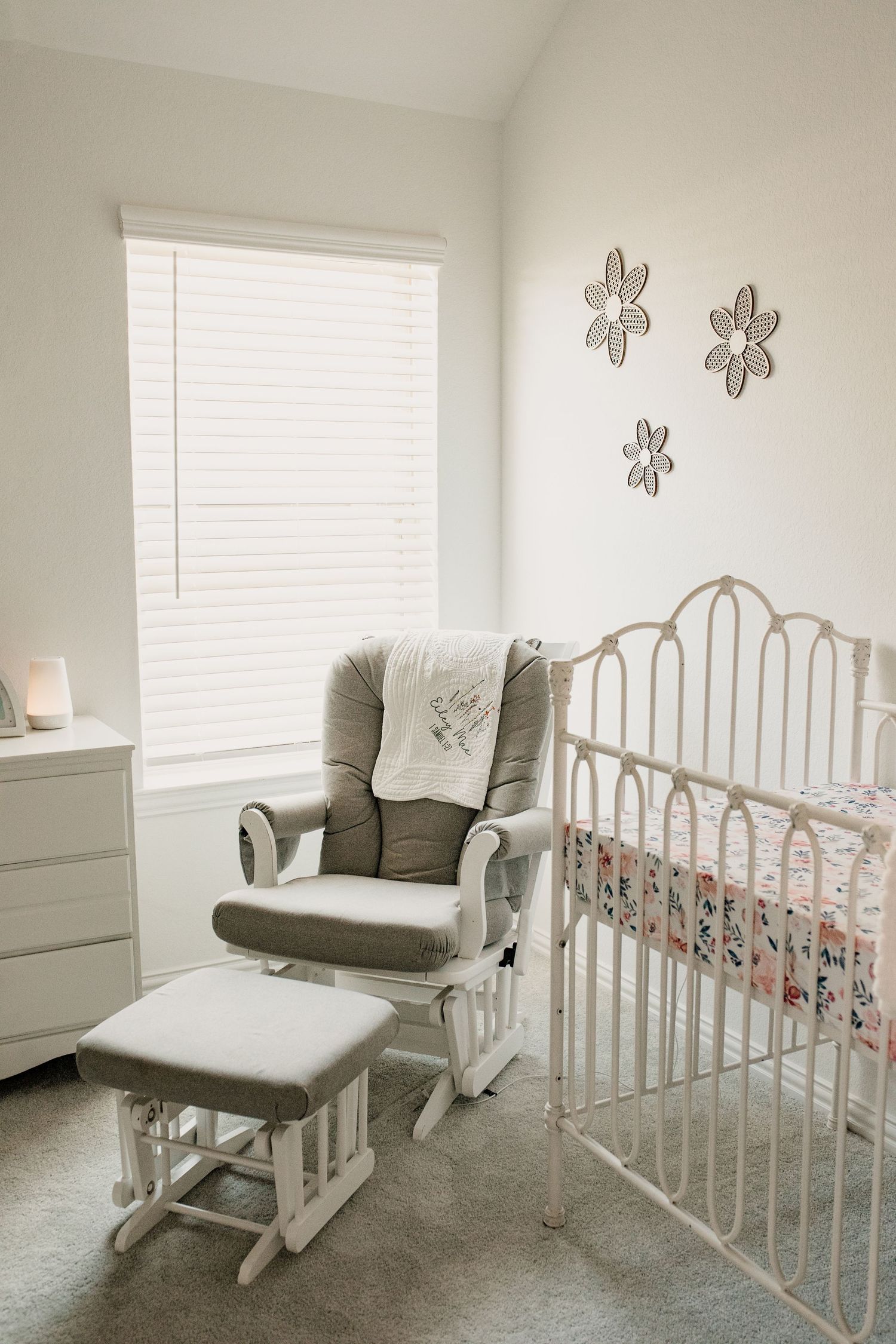 Neutral nursery with white iron crib, gray glider rocking chair, floral wall decor, and soft natural lighting.