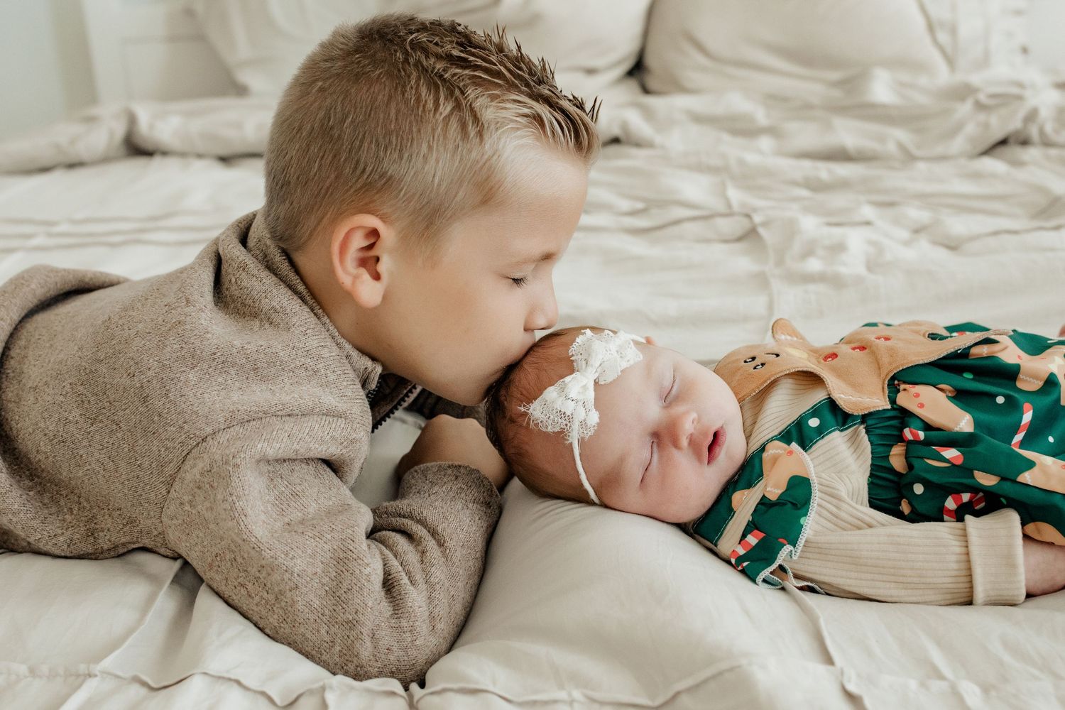 Young boy lovingly kisses his newborn sibling wearing a festive green outfit on a white bed.