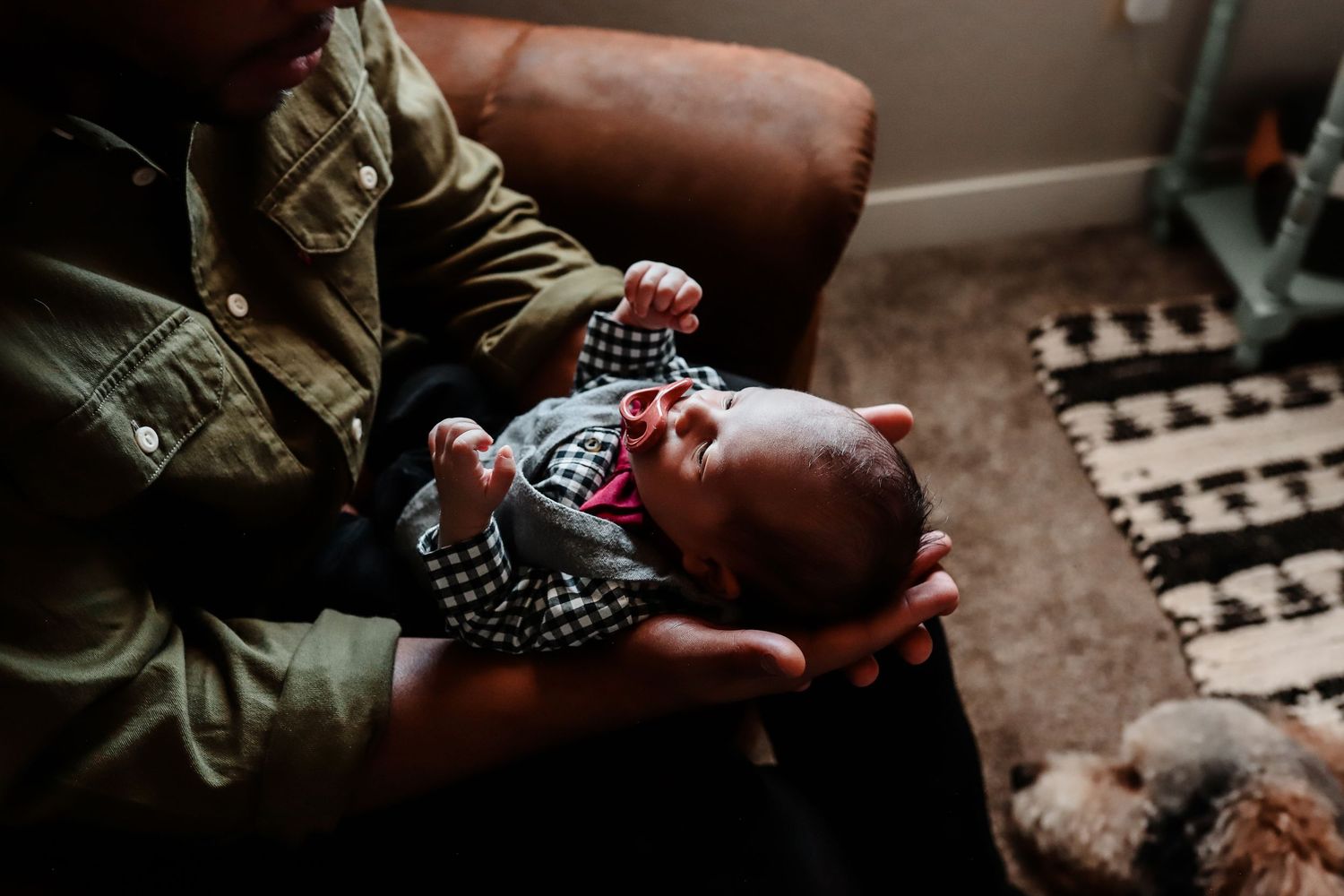 Father holds newborn baby upright in arms beside brown leather chair with family dog visible in background.