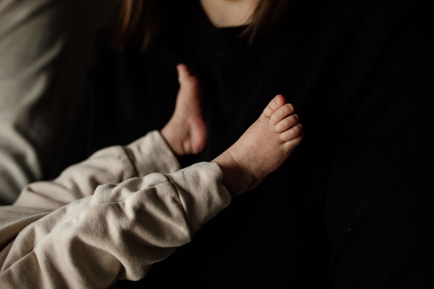 Delicate newborn baby feet resting against a mother wearing a black top in soft, moody lighting.