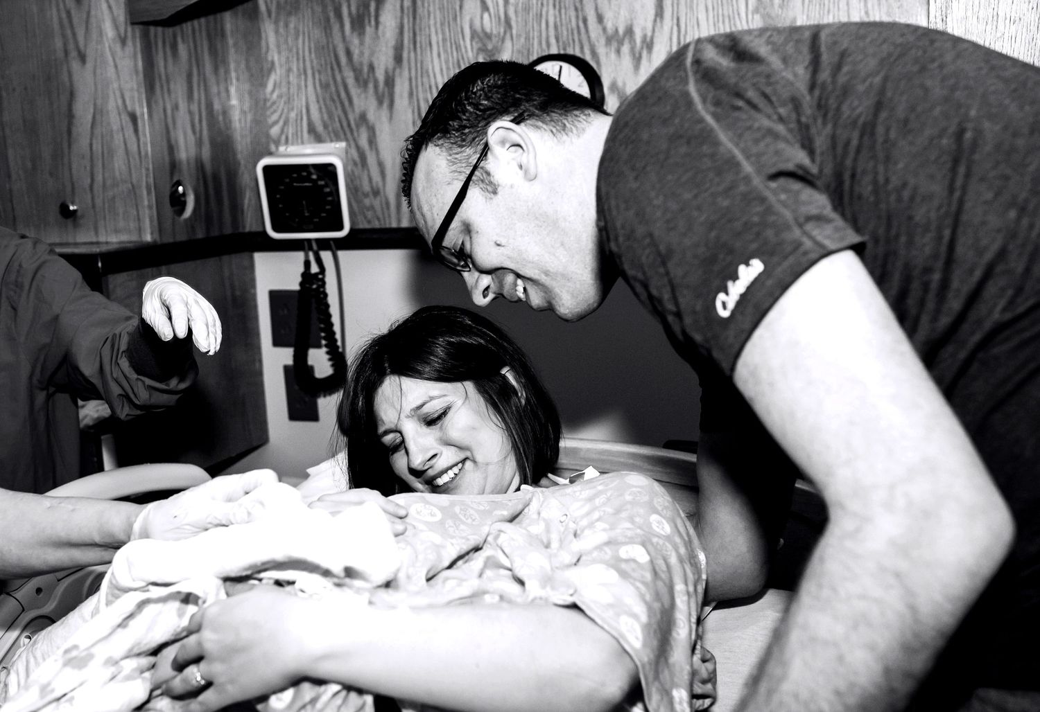 Joyful couple smiles at newborn baby in hospital bed, black and white birth photography moment.