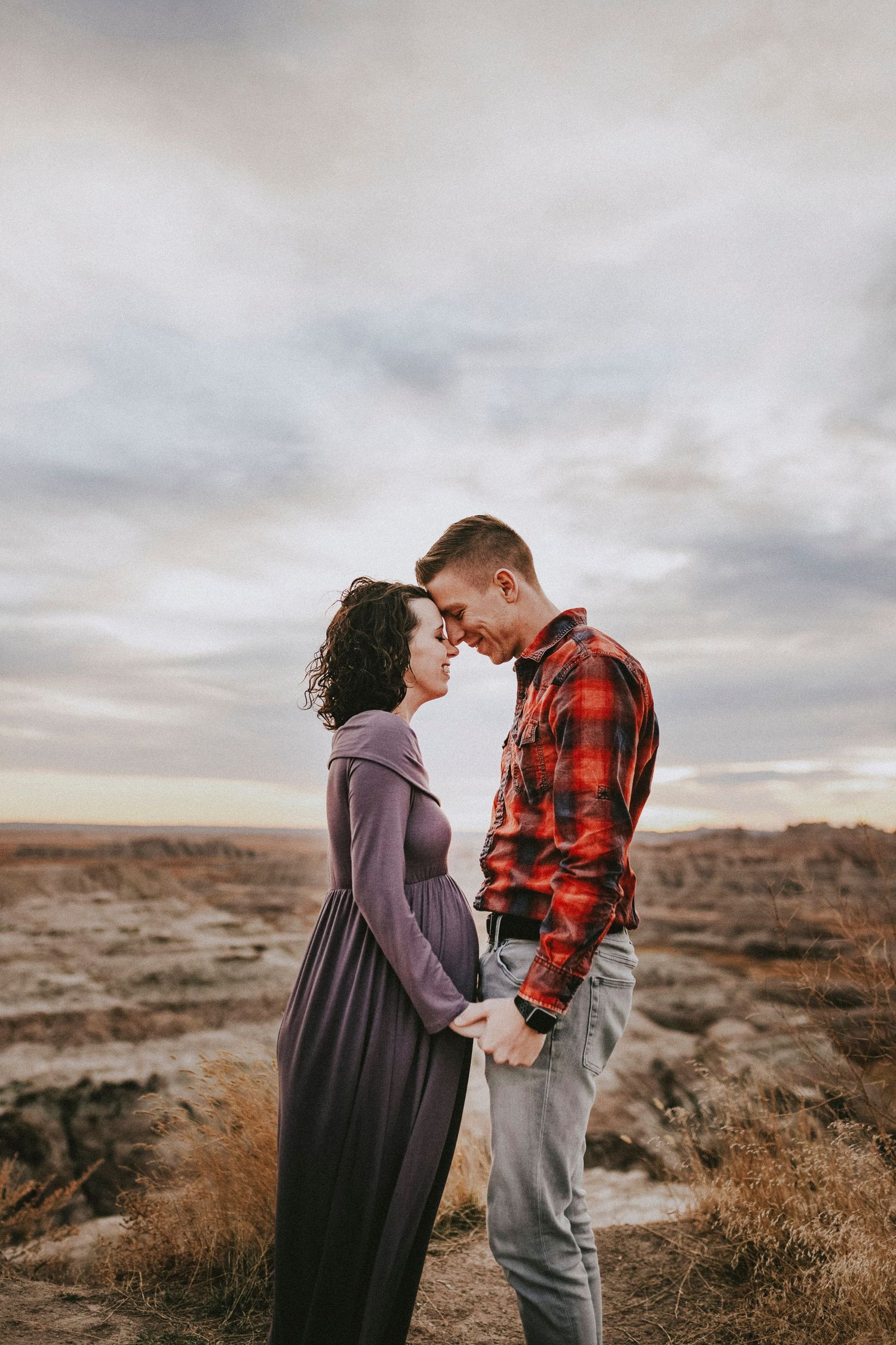 Pregnant couple holding hands facing each other in a dramatic desert landscape under cloudy skies.