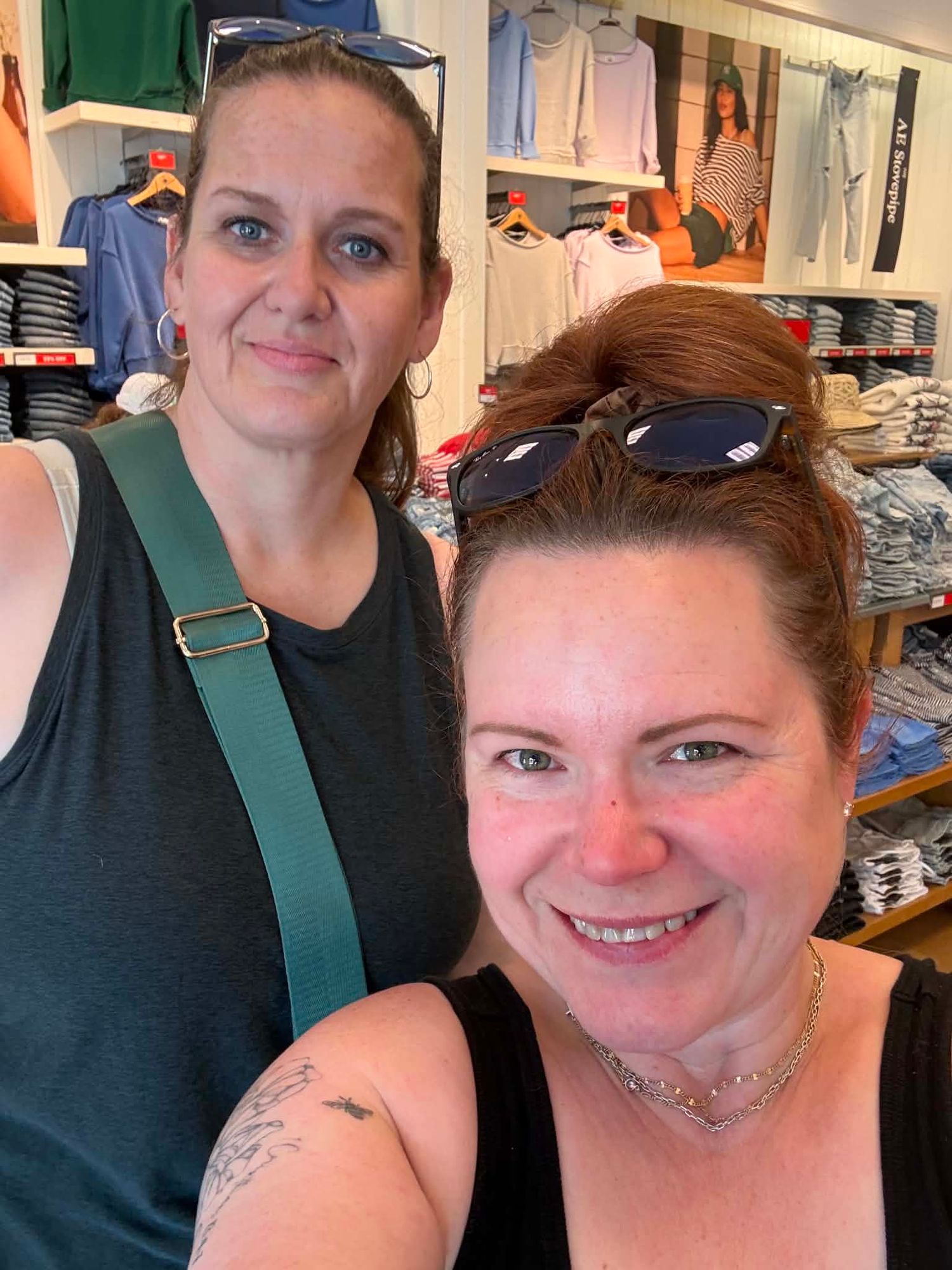 Two women smiling for a selfie inside a clothing store with shelves of folded shirts in the background.